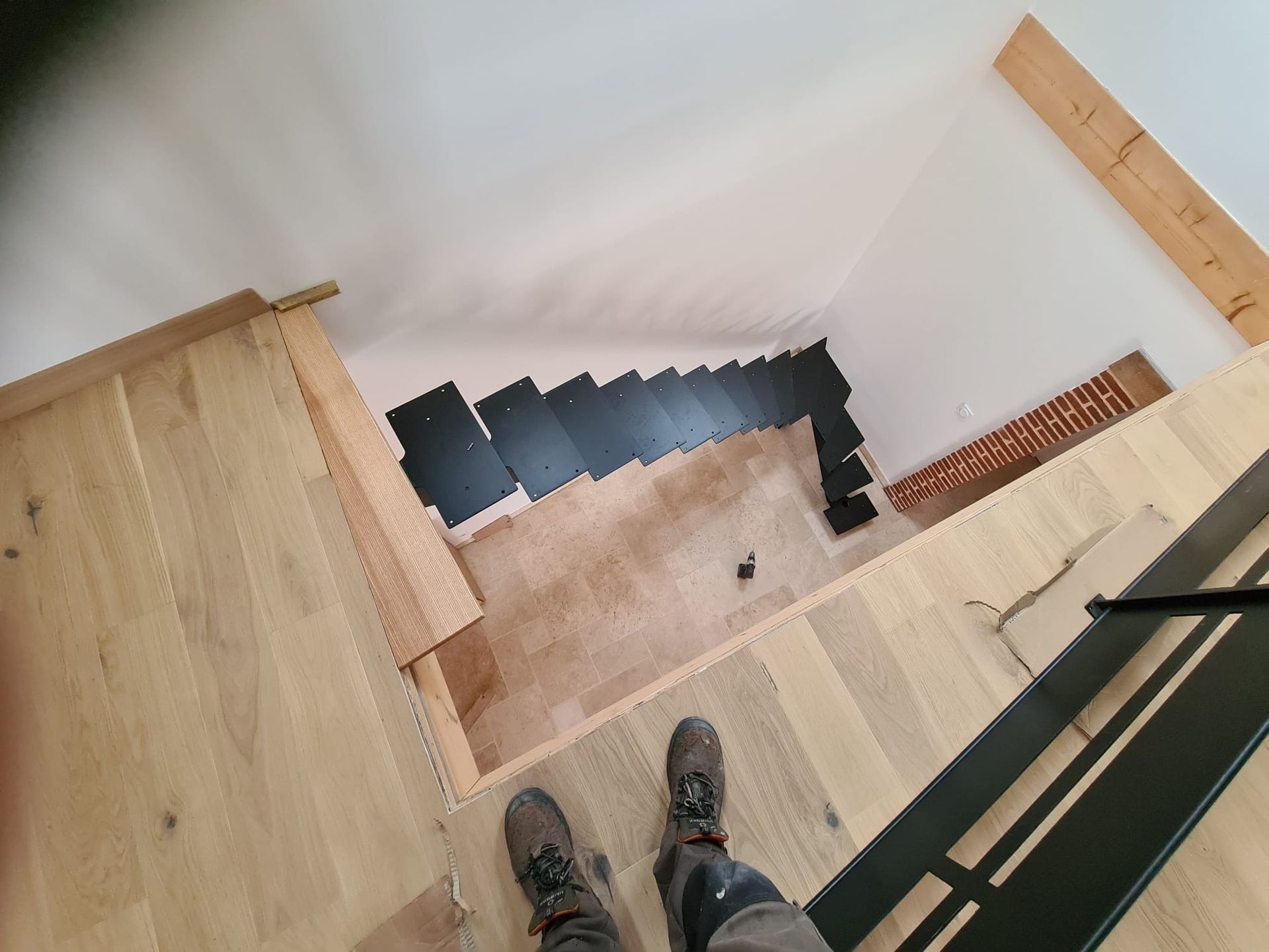 Vue du haut d'un escalier en bois, donnant sur des marches noires et une paire de chaussures sur le palier.