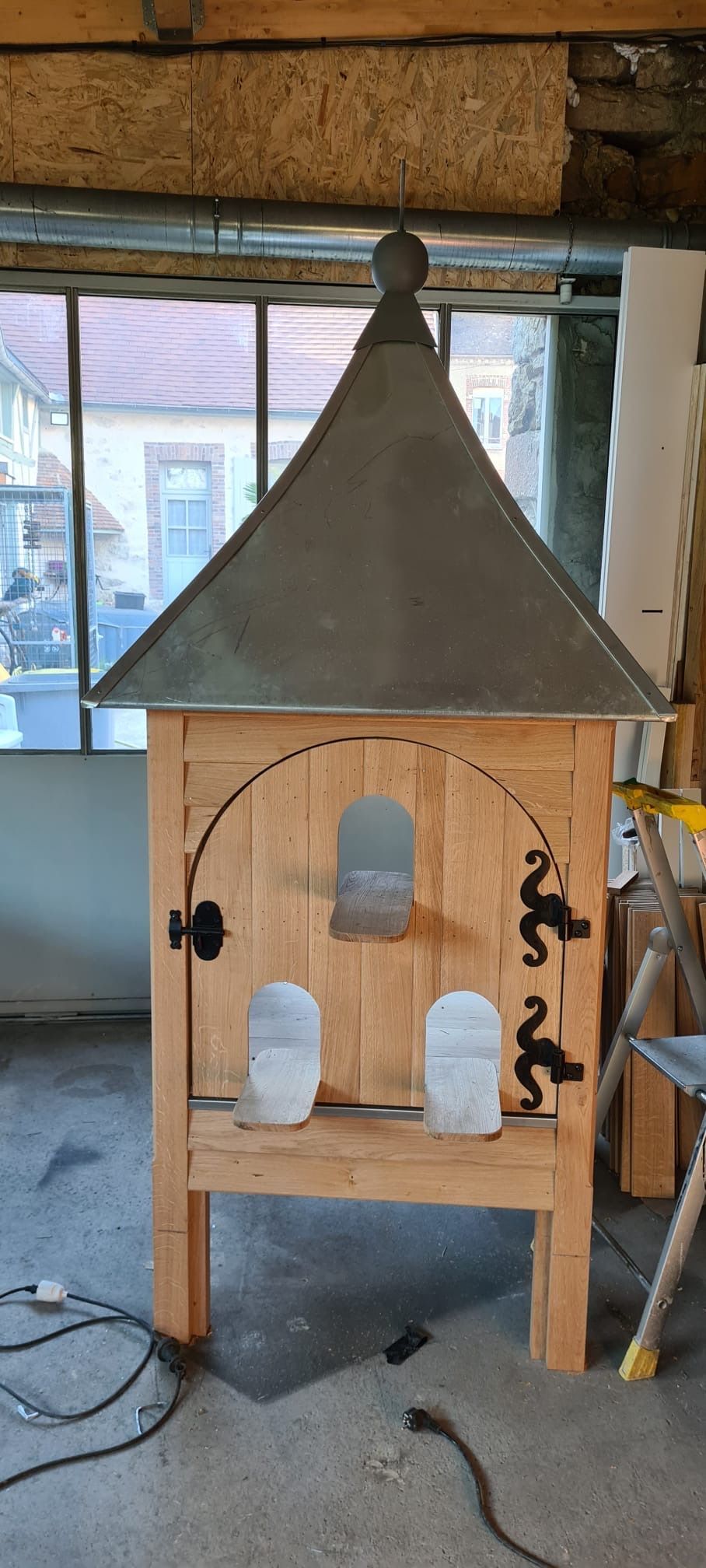 Petite mangeoire à oiseaux en bois avec un toit métallique pointu, suspendue dans un atelier.