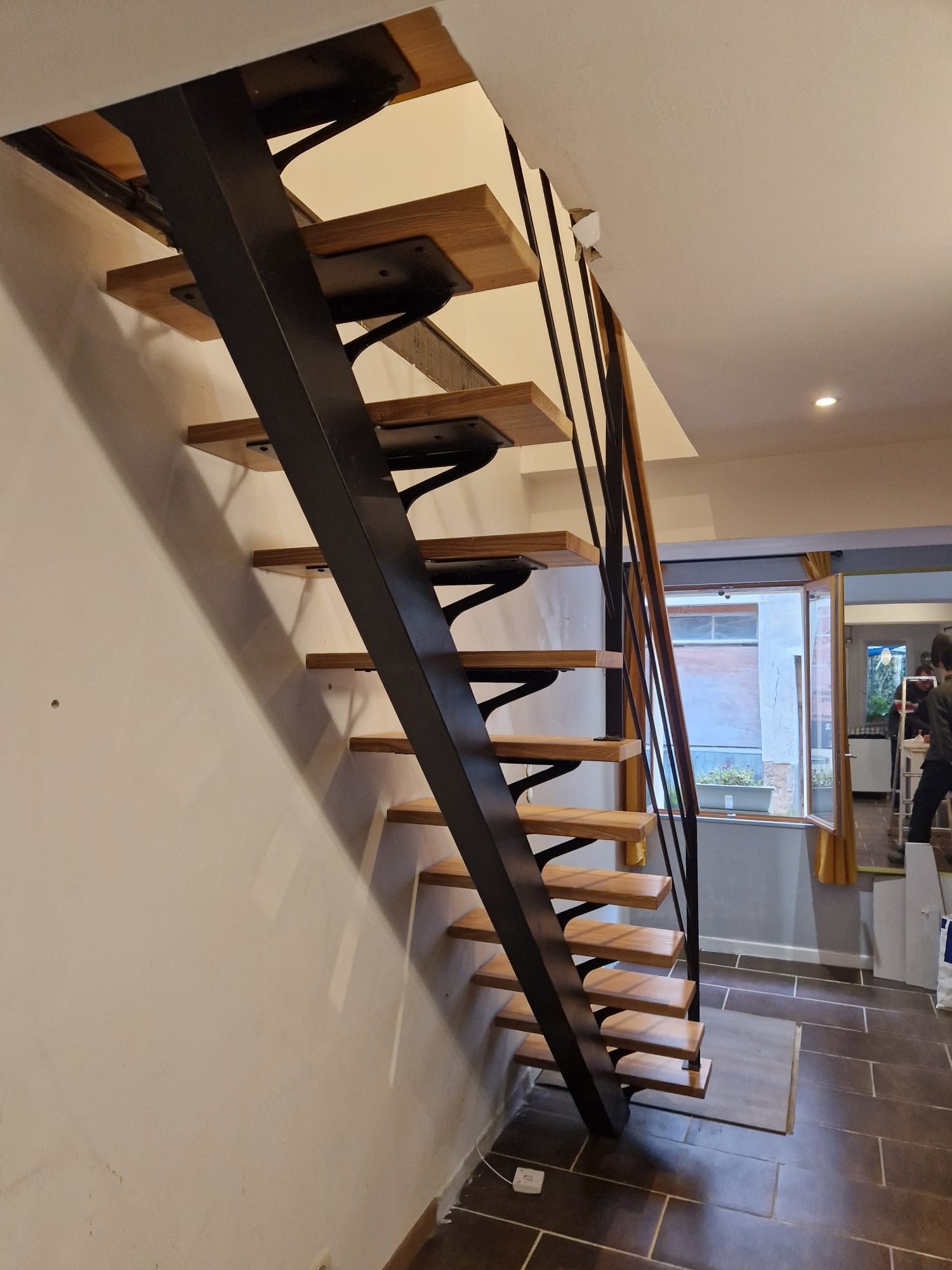 Escalier suspendu dans un intérieur moderne avec marches en bois et supports en métal noir, menant à un couloir lumineux