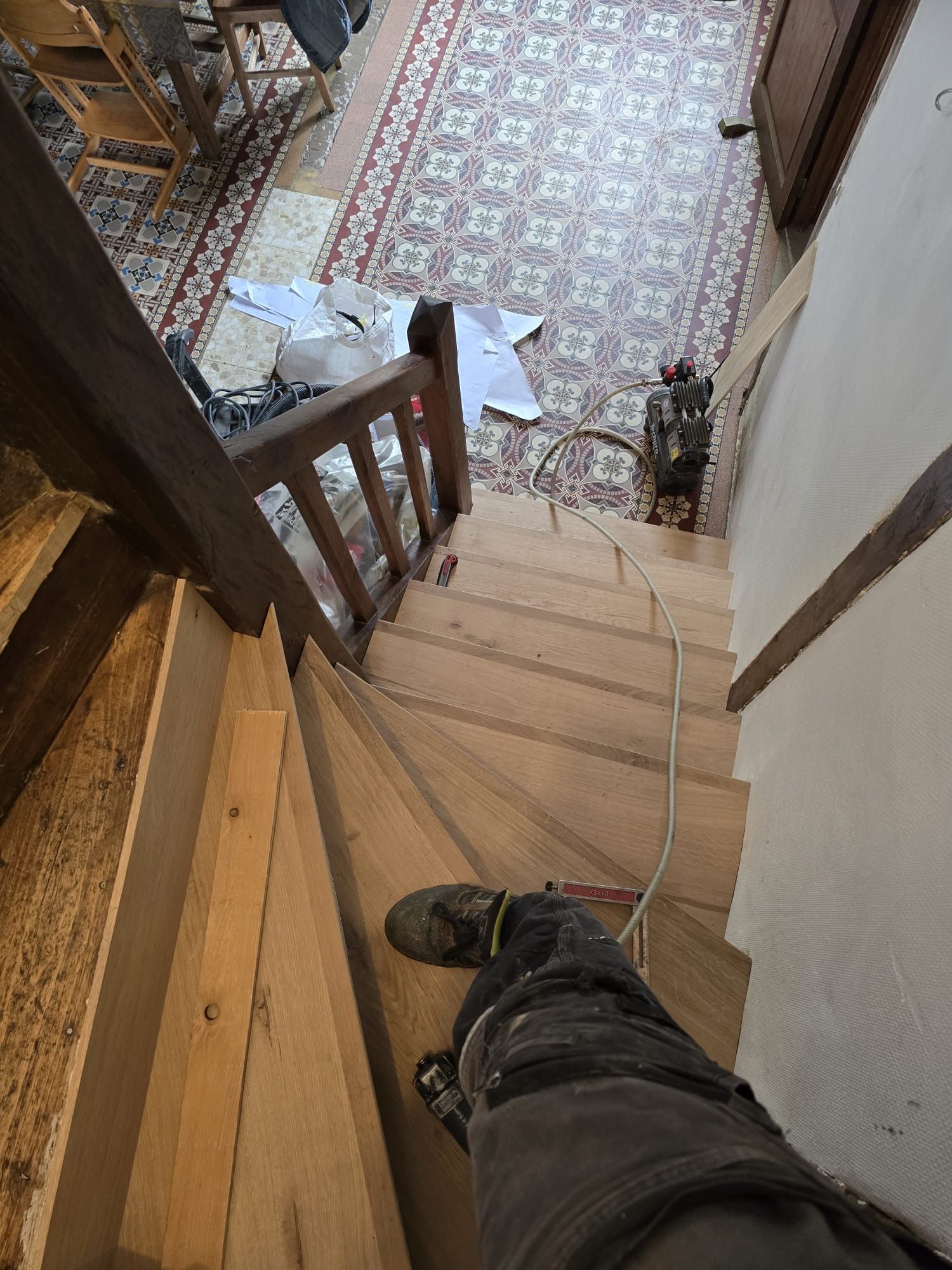 Vue plongeante sur un escalier en bois donnant sur un couloir carrelé, avec la jambe d'une personne visible au premier plan.