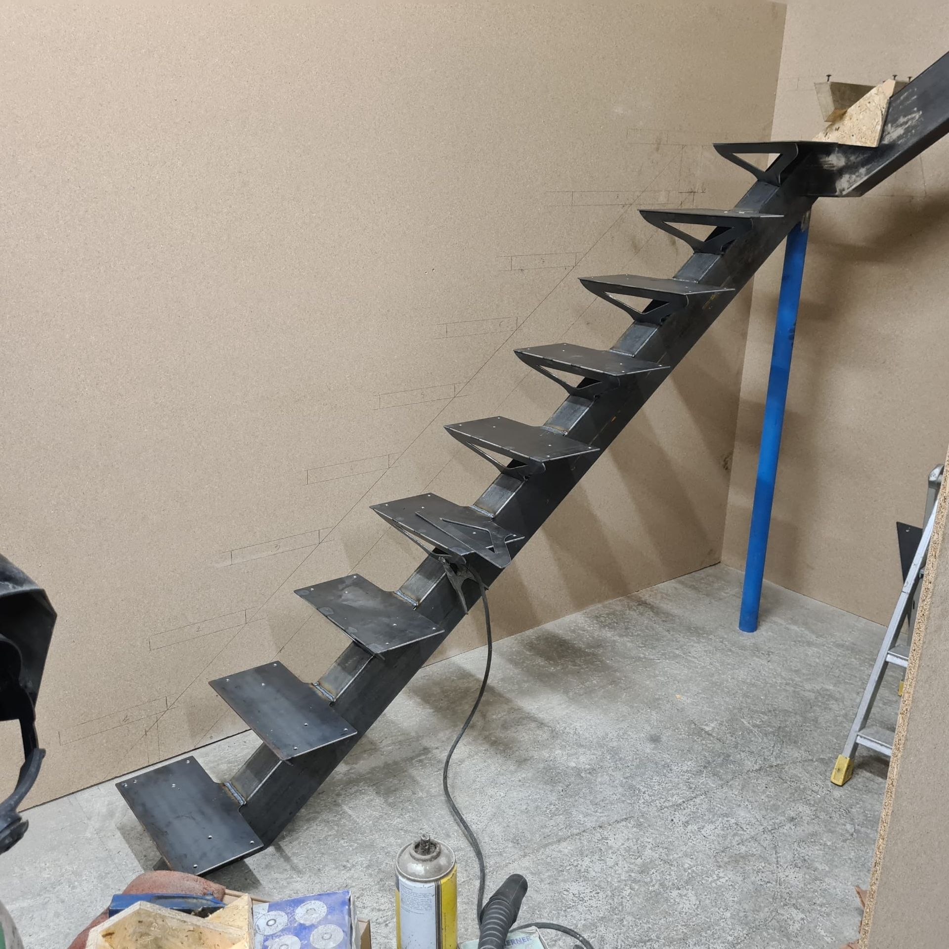 Escalier en métal noir en construction, soutenu par un poteau bleu dans un atelier.