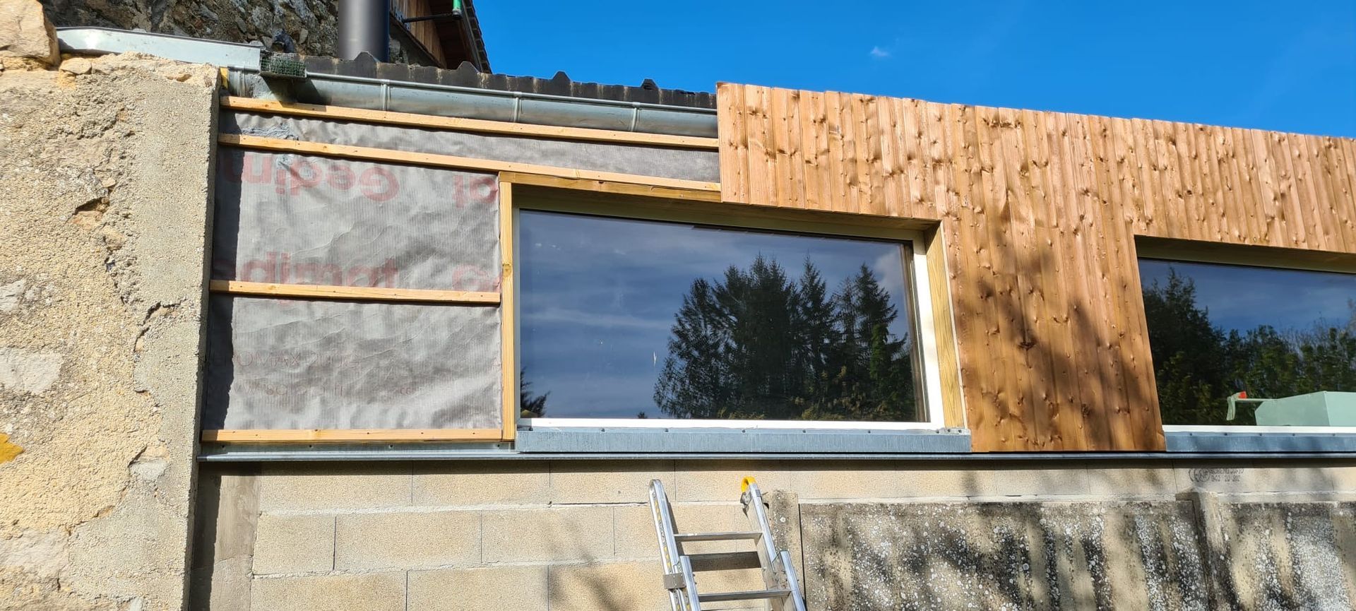 Mur extérieur partiellement fini avec revêtement en pierre et en bois, fenêtres et une échelle en dessous