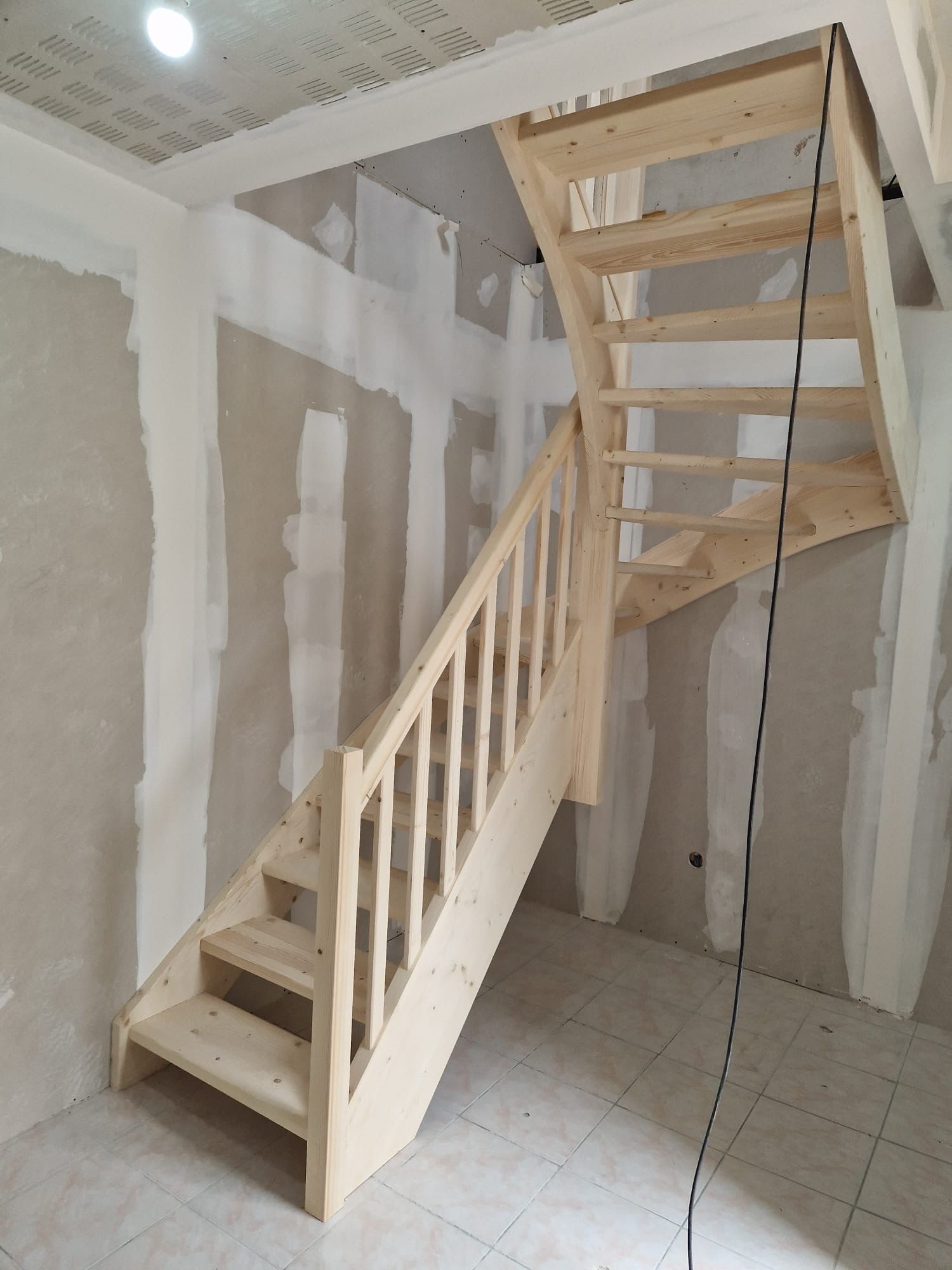 Escalier en bois en construction dans une pièce intérieure inachevée, avec cloisons sèches et plafond apparent.