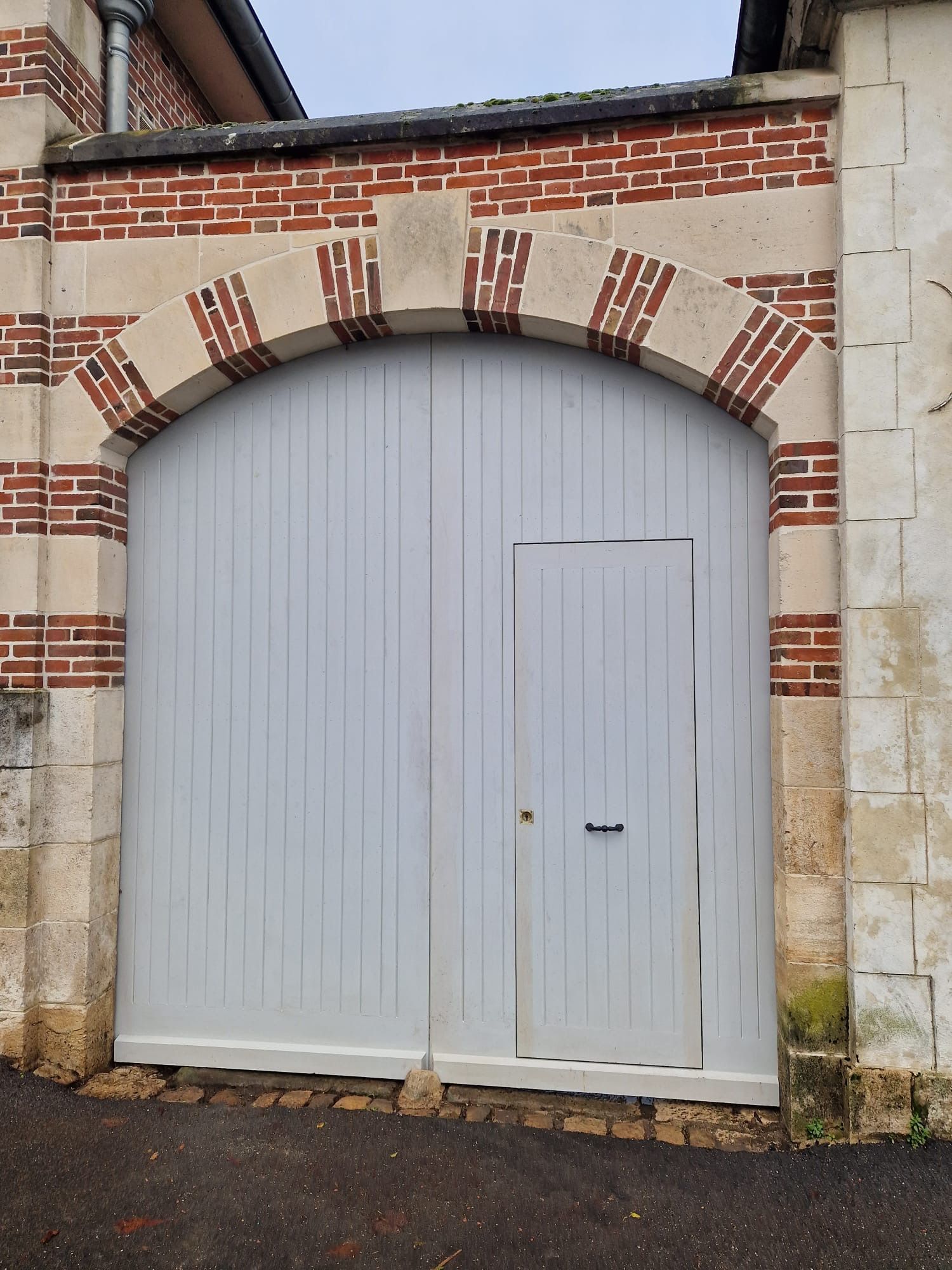 Portail double en bois gris arqué, orné de briques, sur une façade en pierre.