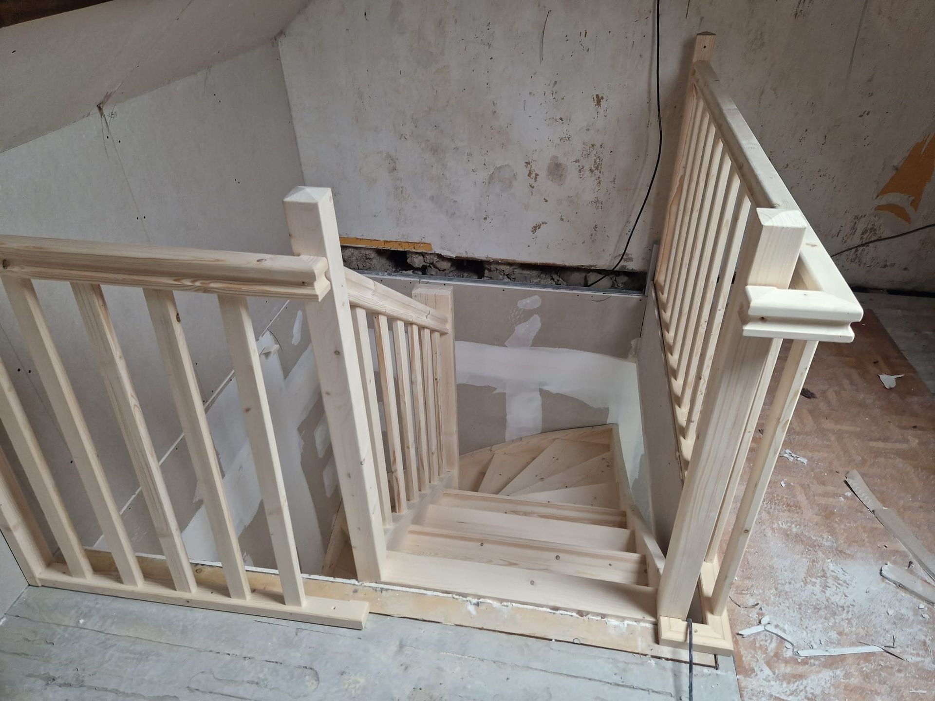 Escalier en bois avec rampe dans un intérieur inachevé, descendant vers une ouverture à l'étage inférieur.