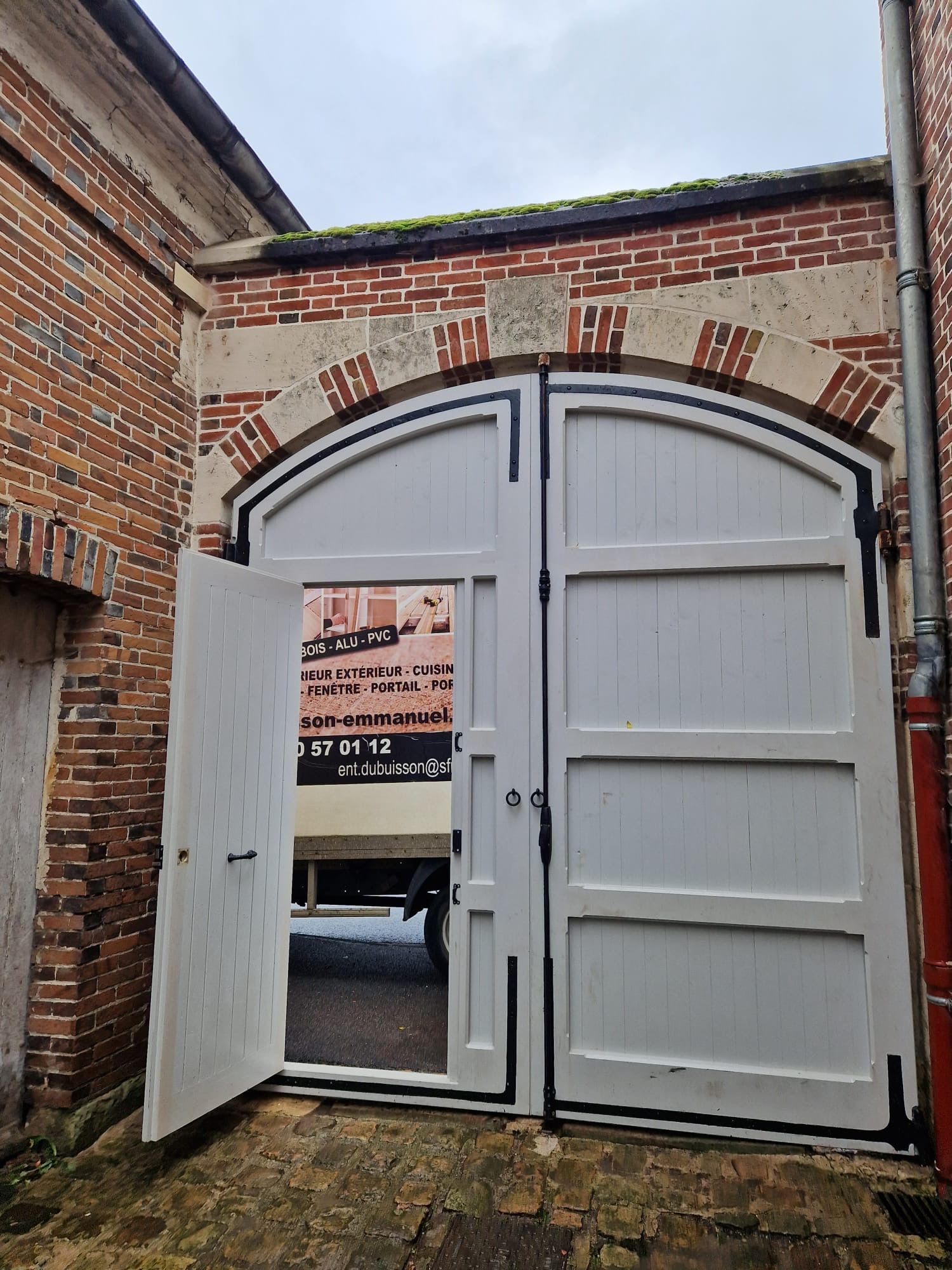 Double porte cintrée blanche ouverte dans une cour en briques, avec une enseigne visible à travers l'embrasure.