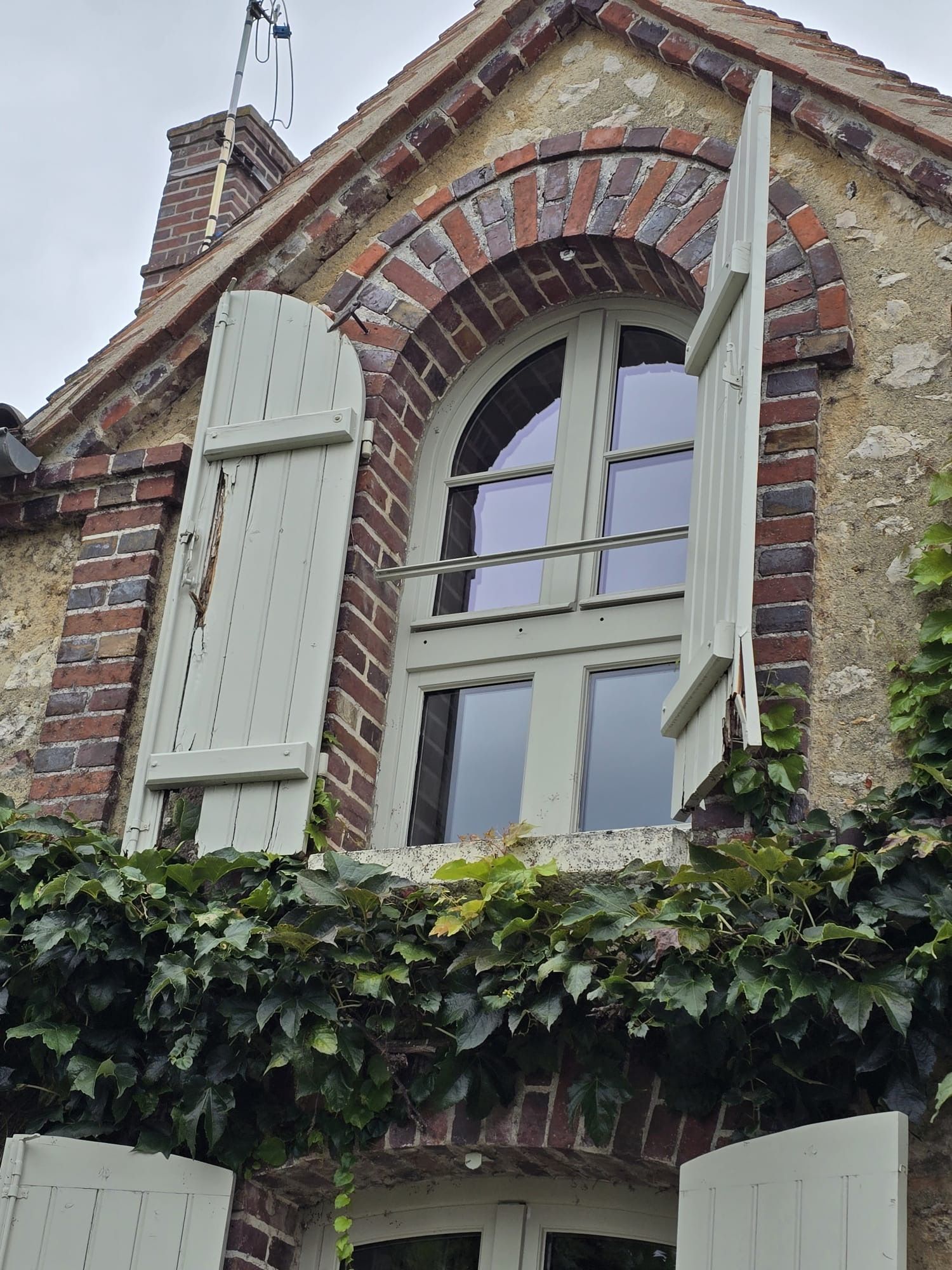 Façade d'une vieille maison en briques avec une fenêtre cintrée, des volets clairs et du lierre en dessous.