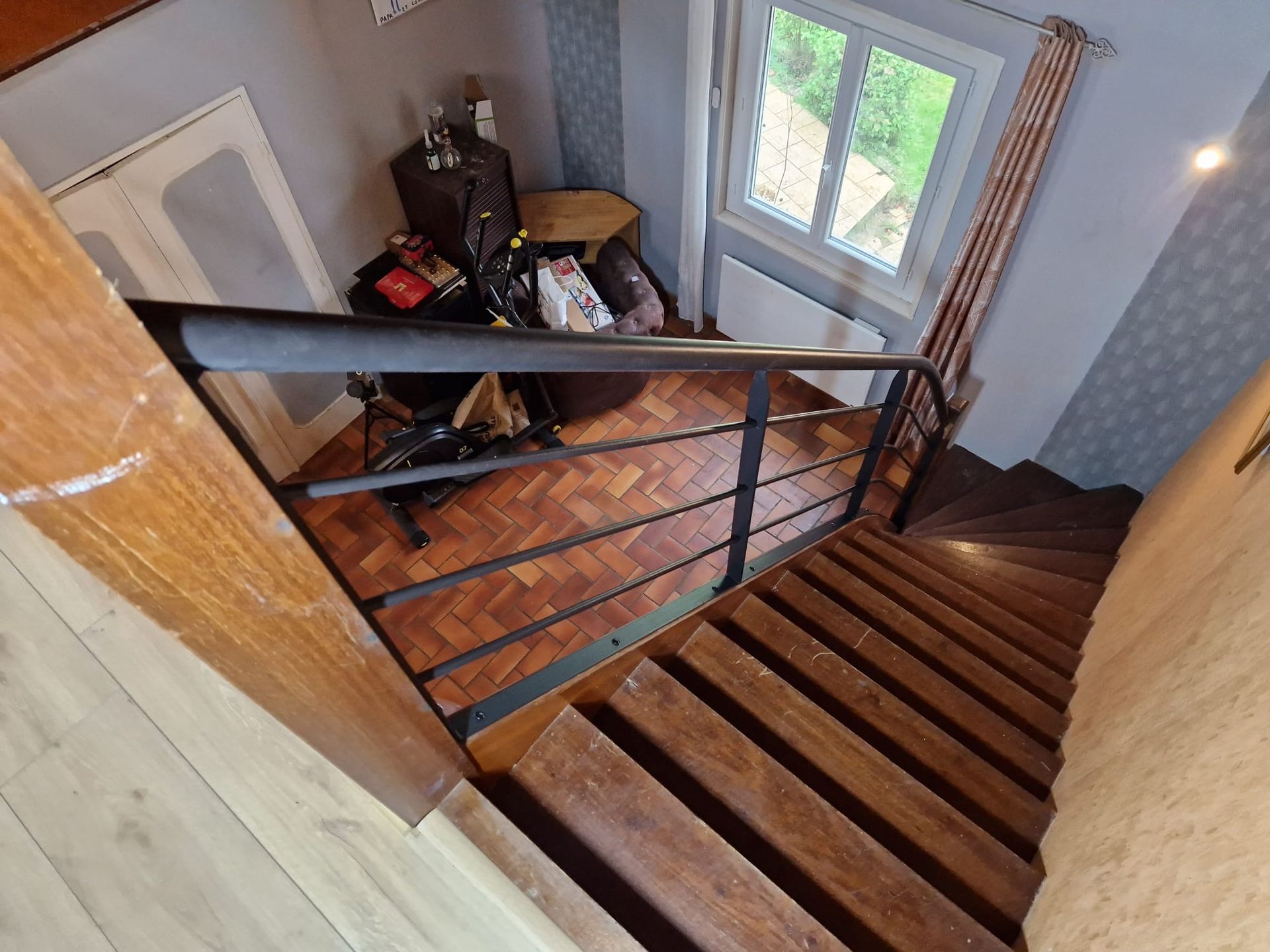 Escalier avec marches en bois, rampe noire et petit palier avec des sacs près d'une fenêtre.