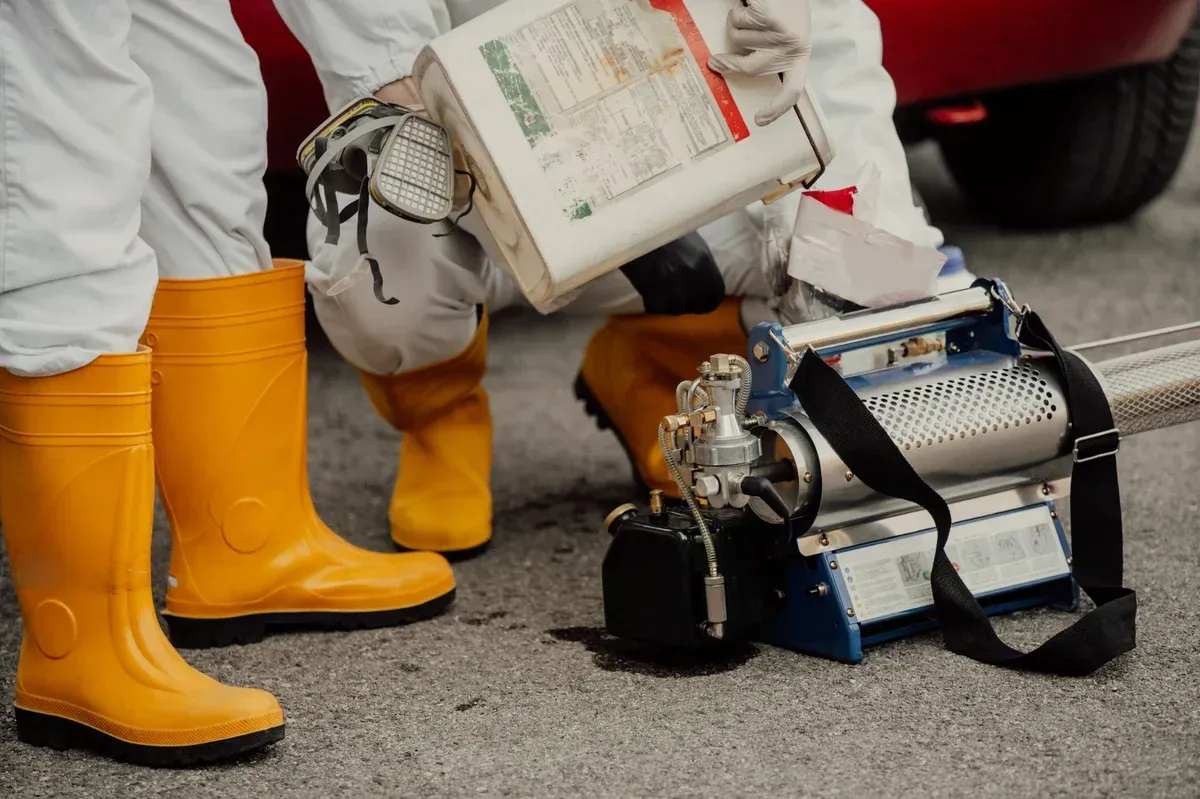 Persona con equipo de protección llenando una máquina nebulizadora azul y plateada, con botas amarillas visibles.