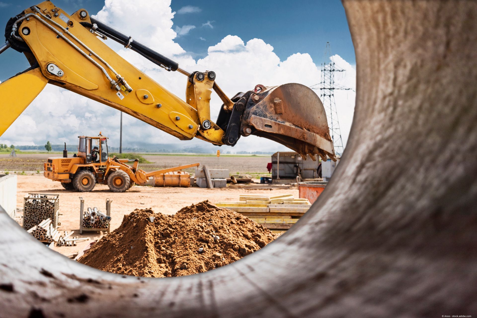 Bagger und Radlader auf einer Baustelle bei Erdarbeiten, aufgenommen durch ein großes Rohr im Vordergrund.