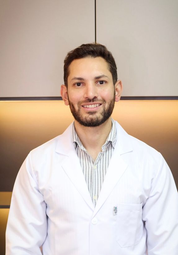 Un hombre sonriente con barba, vestido con una bata blanca y una corbata estampada, posiblemente un médico.
