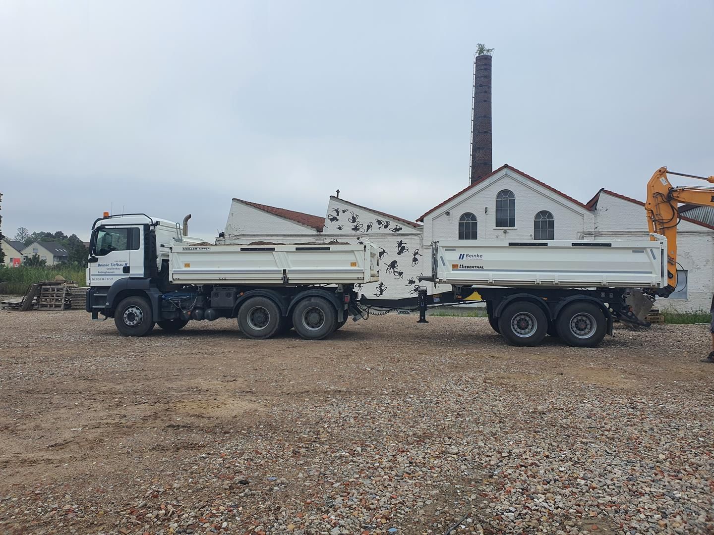 Ein weißer Muldenkipper parkt auf einem Kiesplatz vor einem Gebäude.