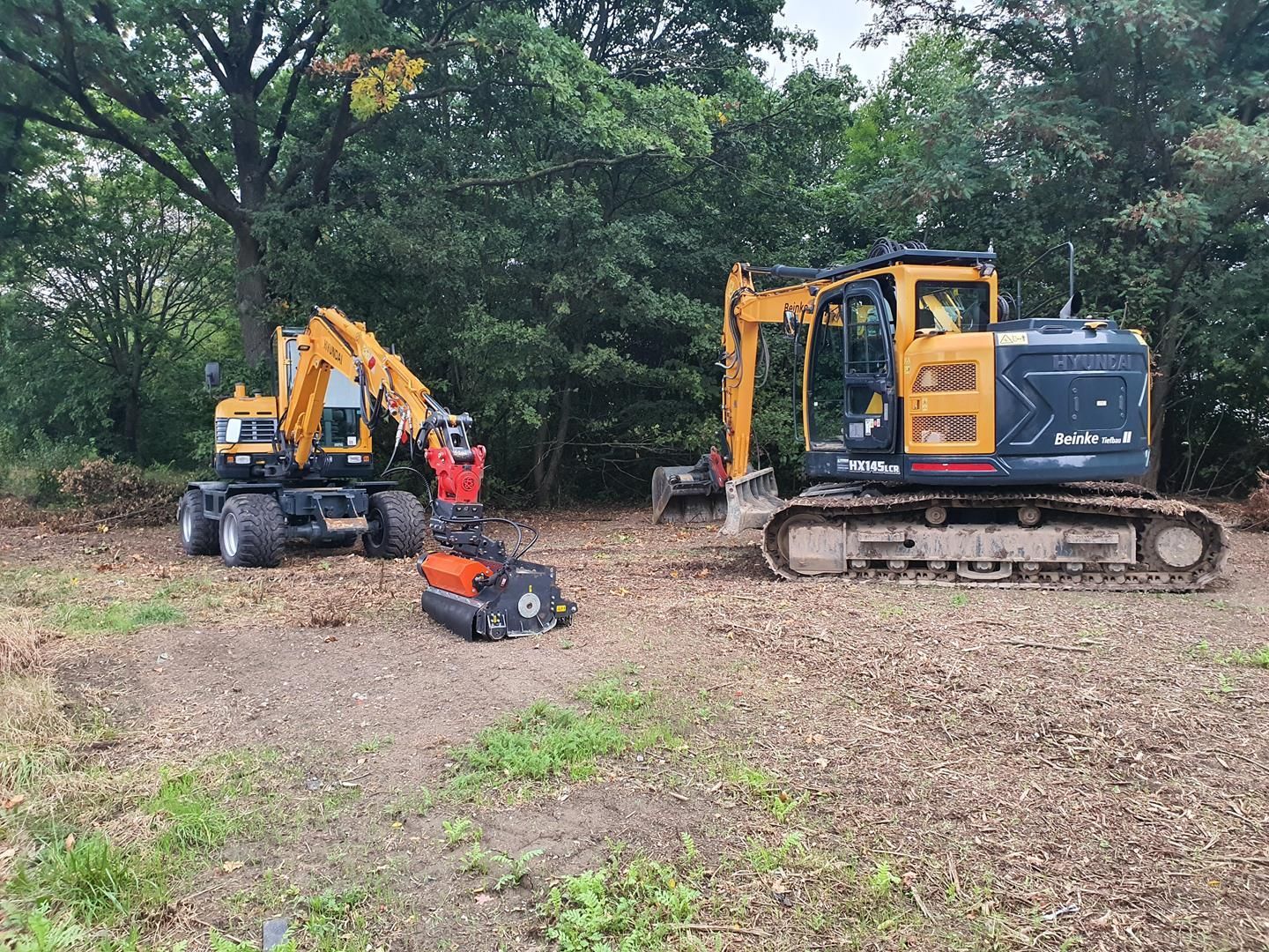Zwei Bagger stehen auf einem Feld mit Bäumen im Hintergrund.