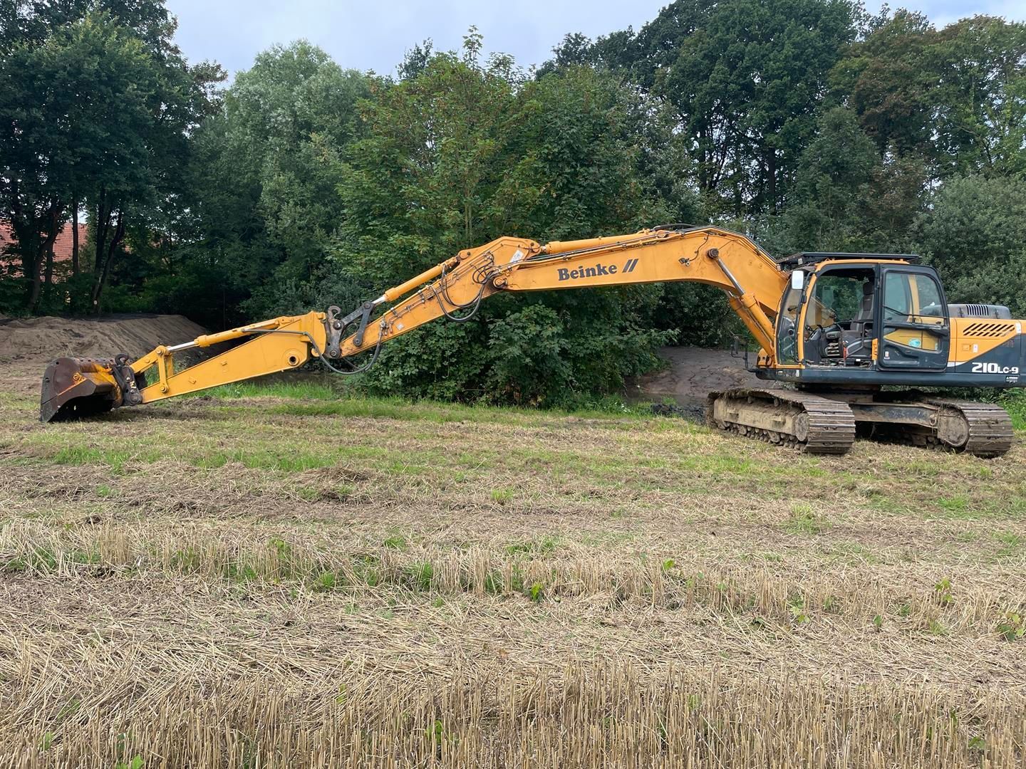 Ein gelber Bagger steht auf einem Feld mit Bäumen im Hintergrund.
