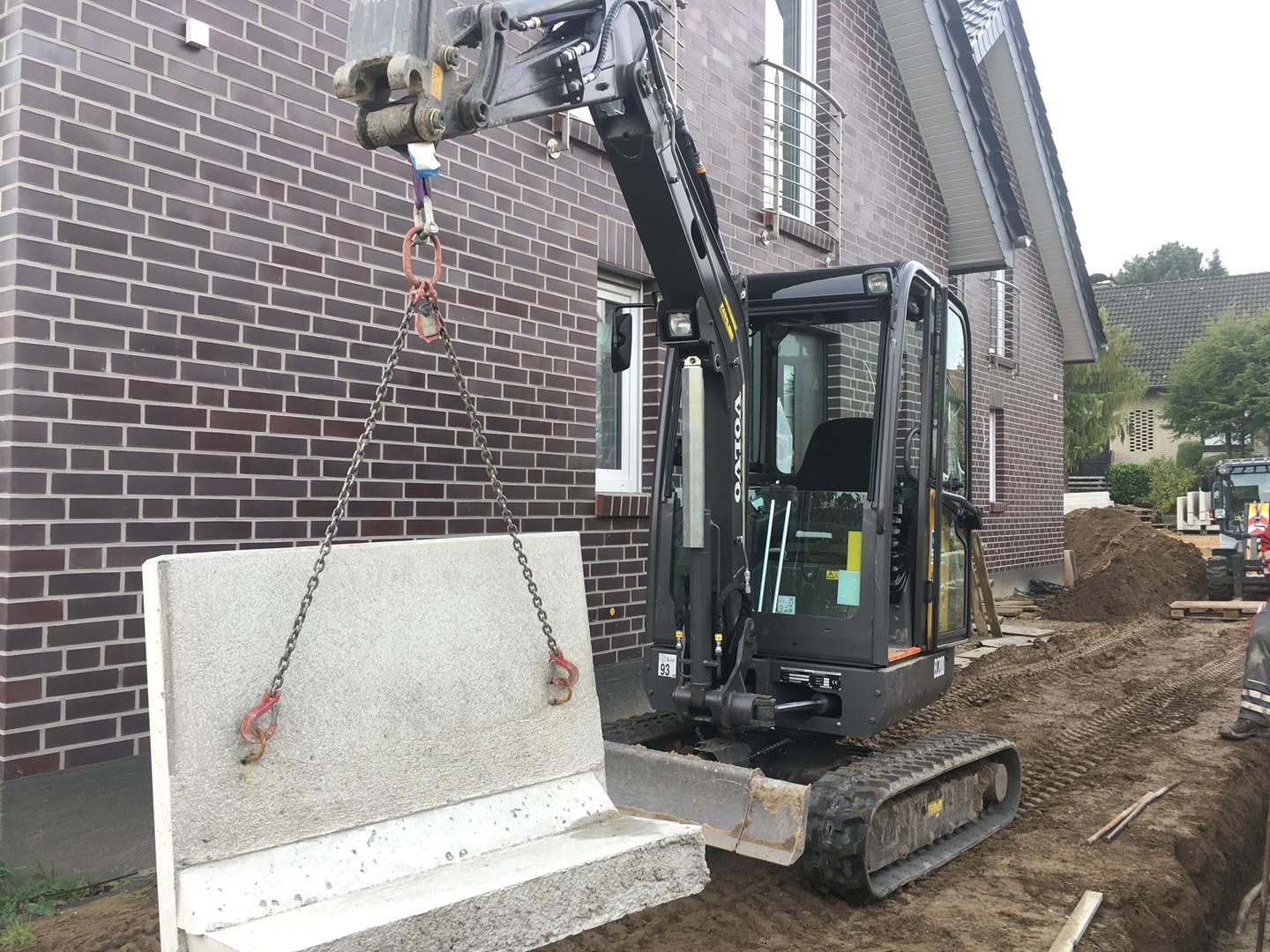 Ein kleiner Bagger hebt eine Betonplatte vor einem Backsteingebäude.