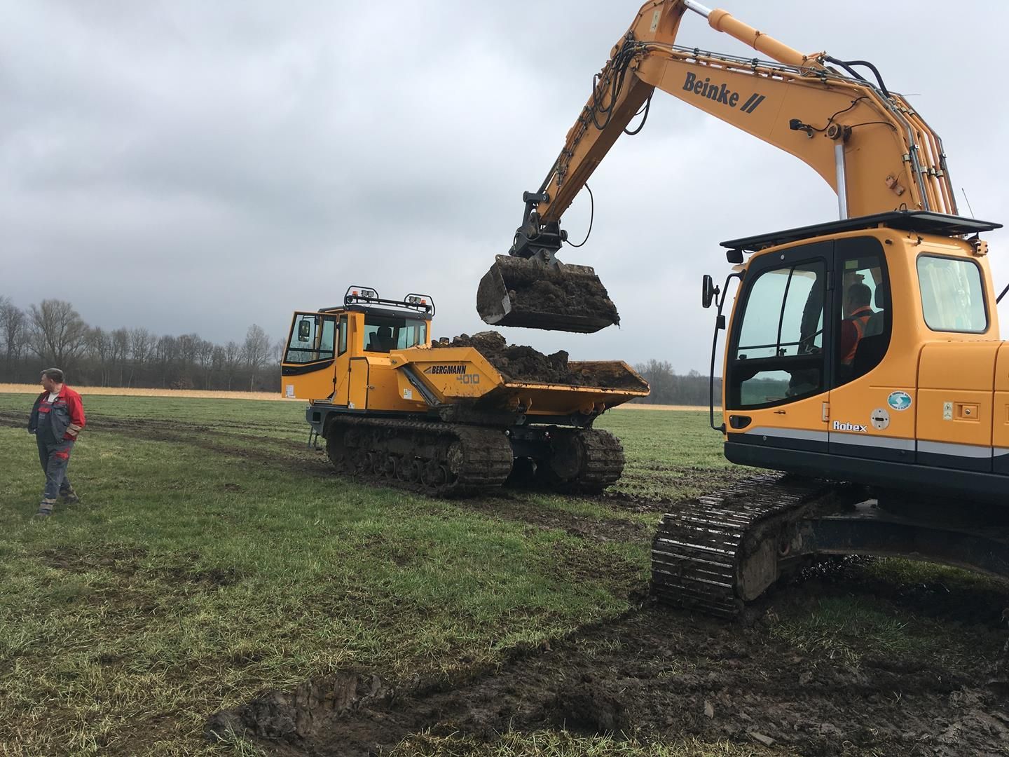 Ein gelber Bagger lädt auf einem Feld Erde in einen Muldenkipper.