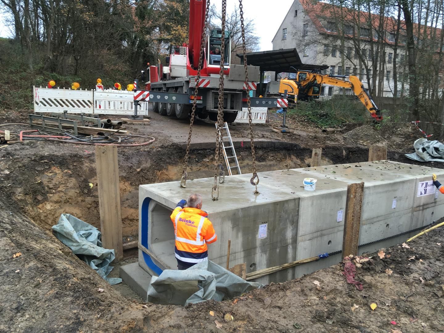 Ein Mann in einer orangefarbenen Jacke steht neben einem großen Betonblock, der von einem Kran angehoben wird.