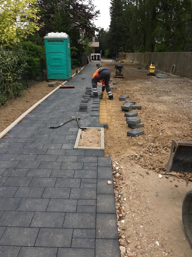 Ein Mann legt Ziegelsteine auf einen Gehweg neben einer grünen mobilen Toilette.