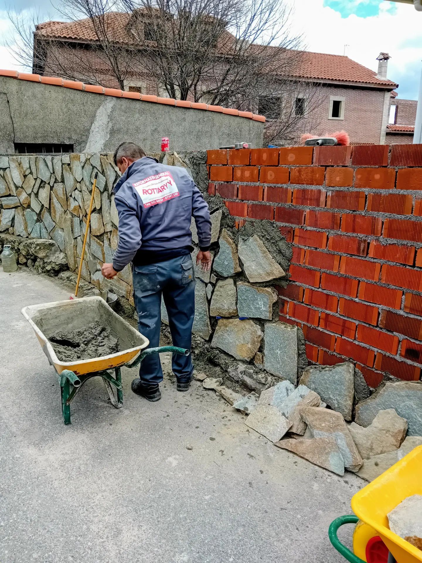 Un hombre empuja una carretilla llena de cemento frente a una pared de ladrillos.