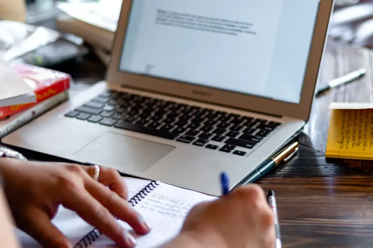 Persona escribiendo en un cuaderno, con una computadora portátil abierta al frente y libros en el fondo.