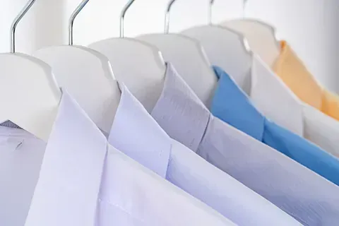Una hilera de camisas de vestir para hombre en tonos blancos, azul claro y amarillo, colgadas en perchas sobre un fondo blanco.