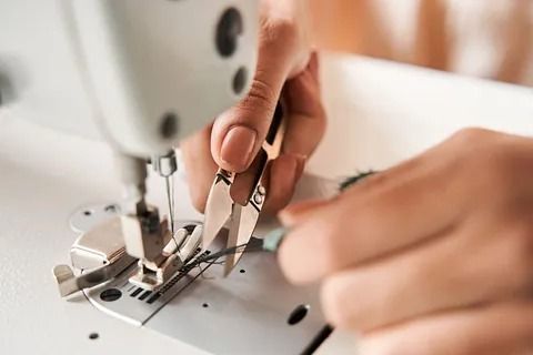 Primer plano de unas manos que utilizan unas pequeñas tijeras metálicas para cortar el hilo que queda debajo del prensatelas de una máquina de coser.