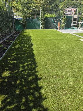 Un gazebo in legno si erge accanto a un rigoglioso giardino verdeggiante con un albero piangente e una recinzione in legno, sullo sfondo di una pineta.