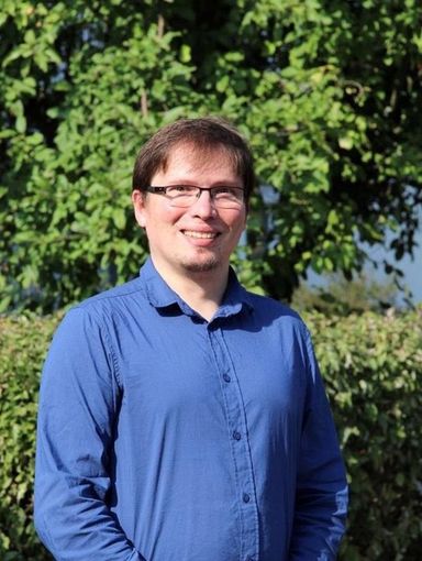Lächelnder Mann mit Brille und blauem Hemd, der im Freien vor grünem Laub steht.