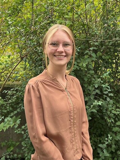 Frau mit blonden Haaren, Brille und braunem Hemd lächelt vor grünem Laub.