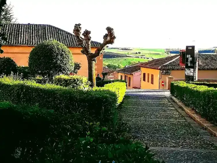 Sendero de piedra junto a setos recortados y casas, con vistas a un luminoso valle rural.