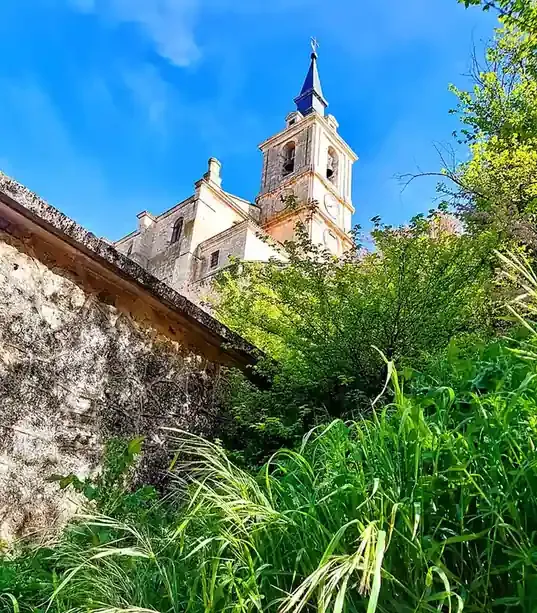 Torre de piedra de una iglesia sobre un follaje verde y un muro desgastado bajo un cielo azul brillante