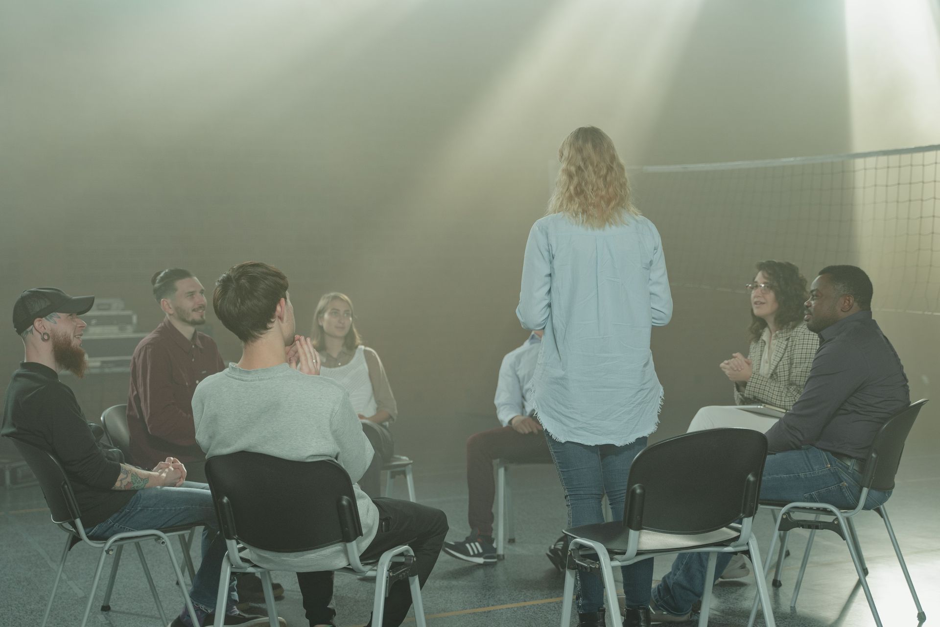 Des personnes assises en cercle écoutent un orateur debout dans une salle de réunion enfumée.