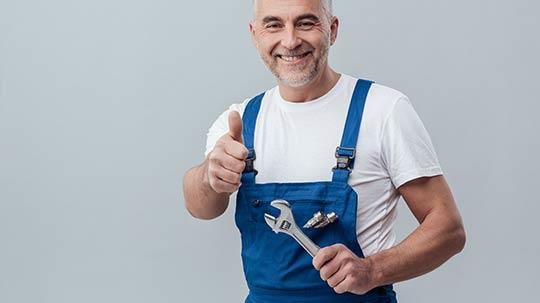 Ein Mann im blauen Overall hält einen Schraubenschlüssel und streckt den Daumen nach oben.