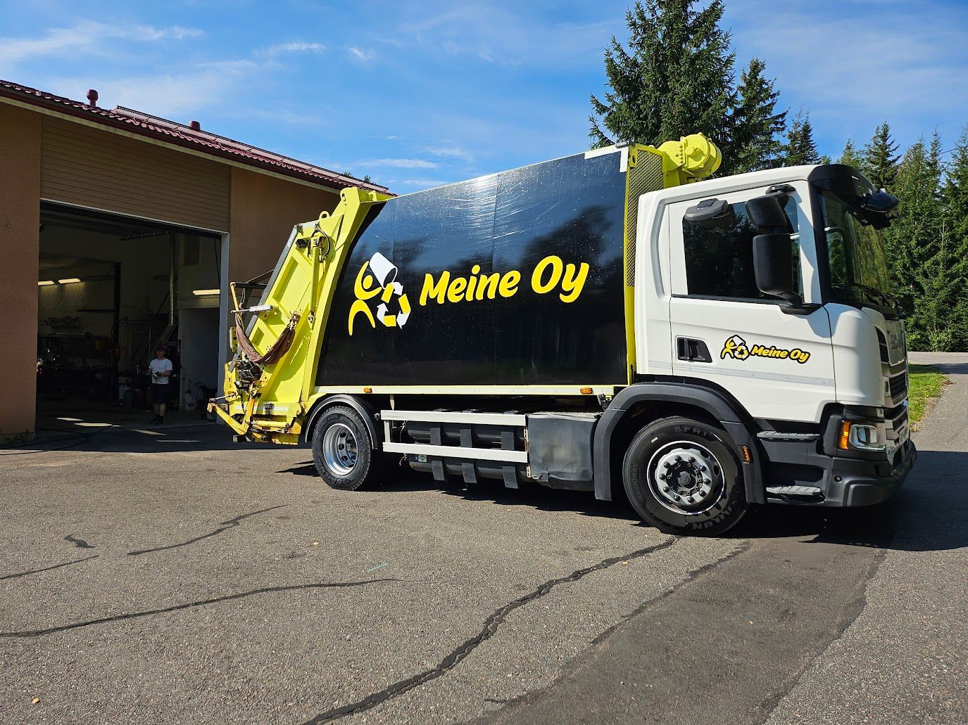 Valkoinen ja musta Meine Oy:n jäteauto pysäköitynä rakennuksen ulkopuolelle asfaltille.