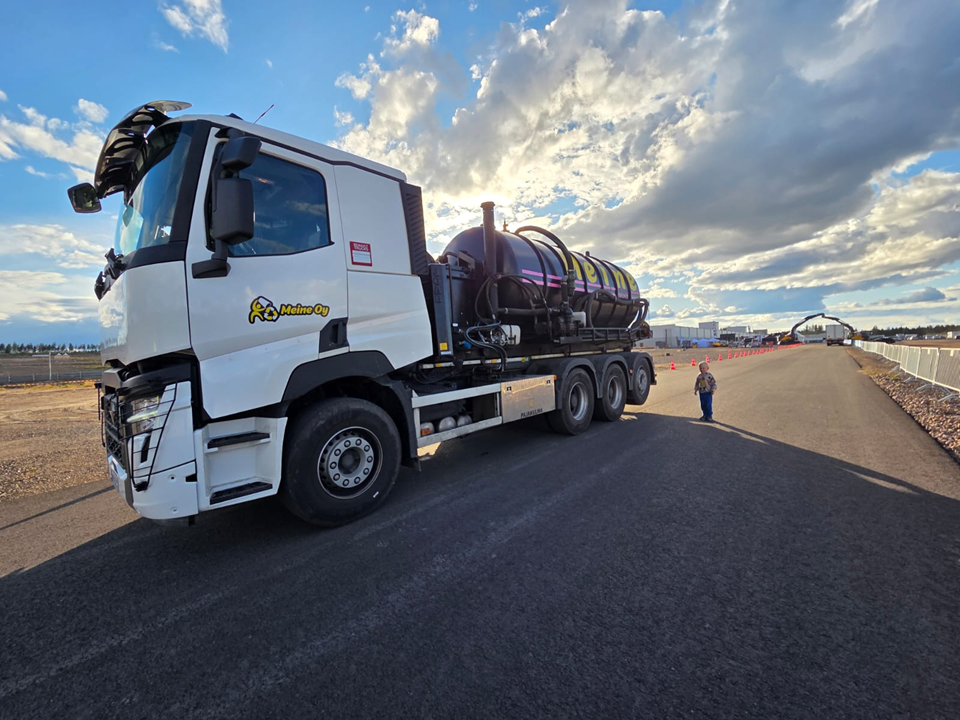 Valkoinen kuorma-auto mustalla säiliöllä pysäköitynä asfalttitielle, henkilö seisoo lähellä, sininen taivas.