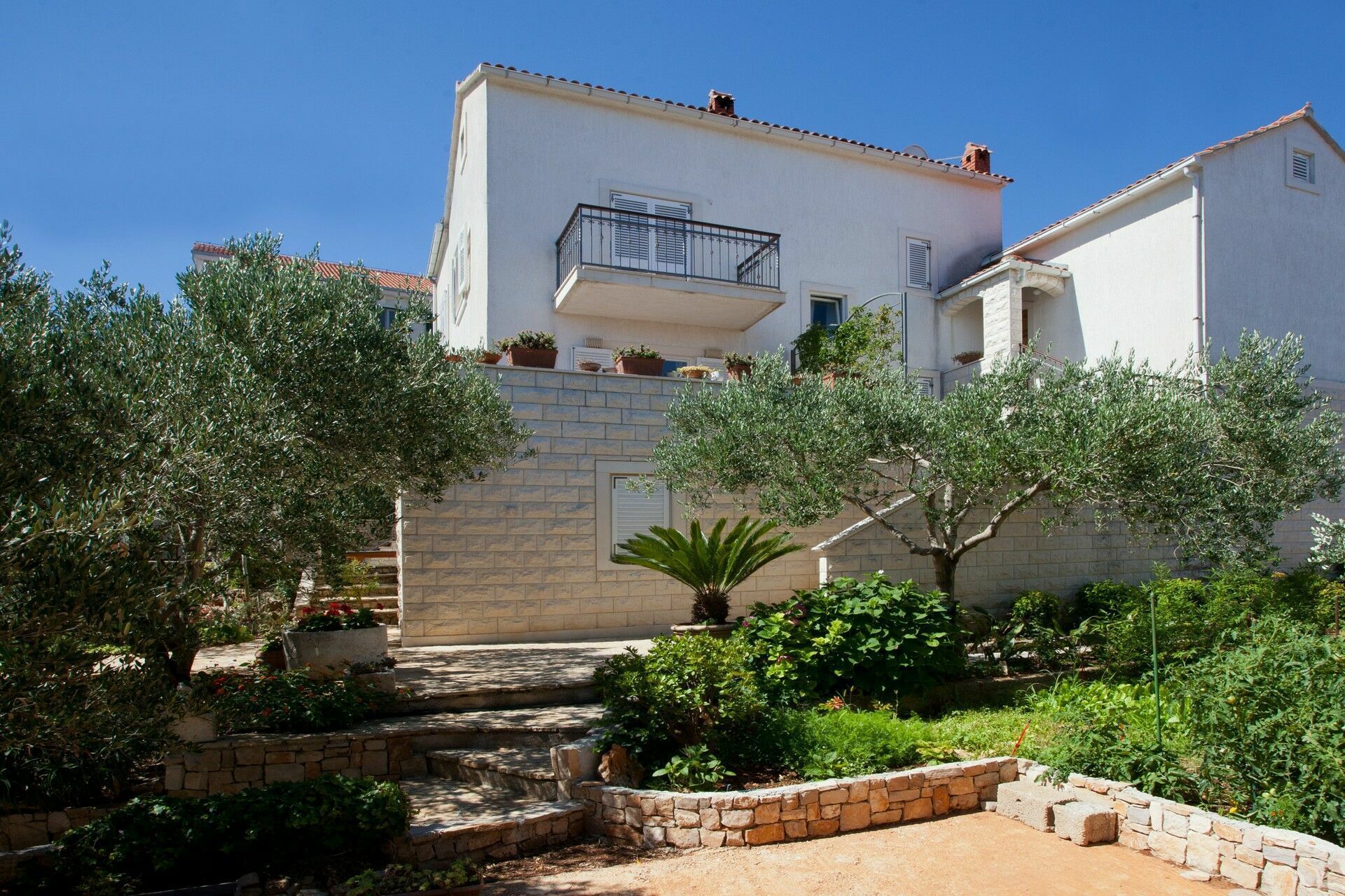 A white villa with a balcony is surrounded by trees and bushes, located in Supetar, Brac Island, Croatia