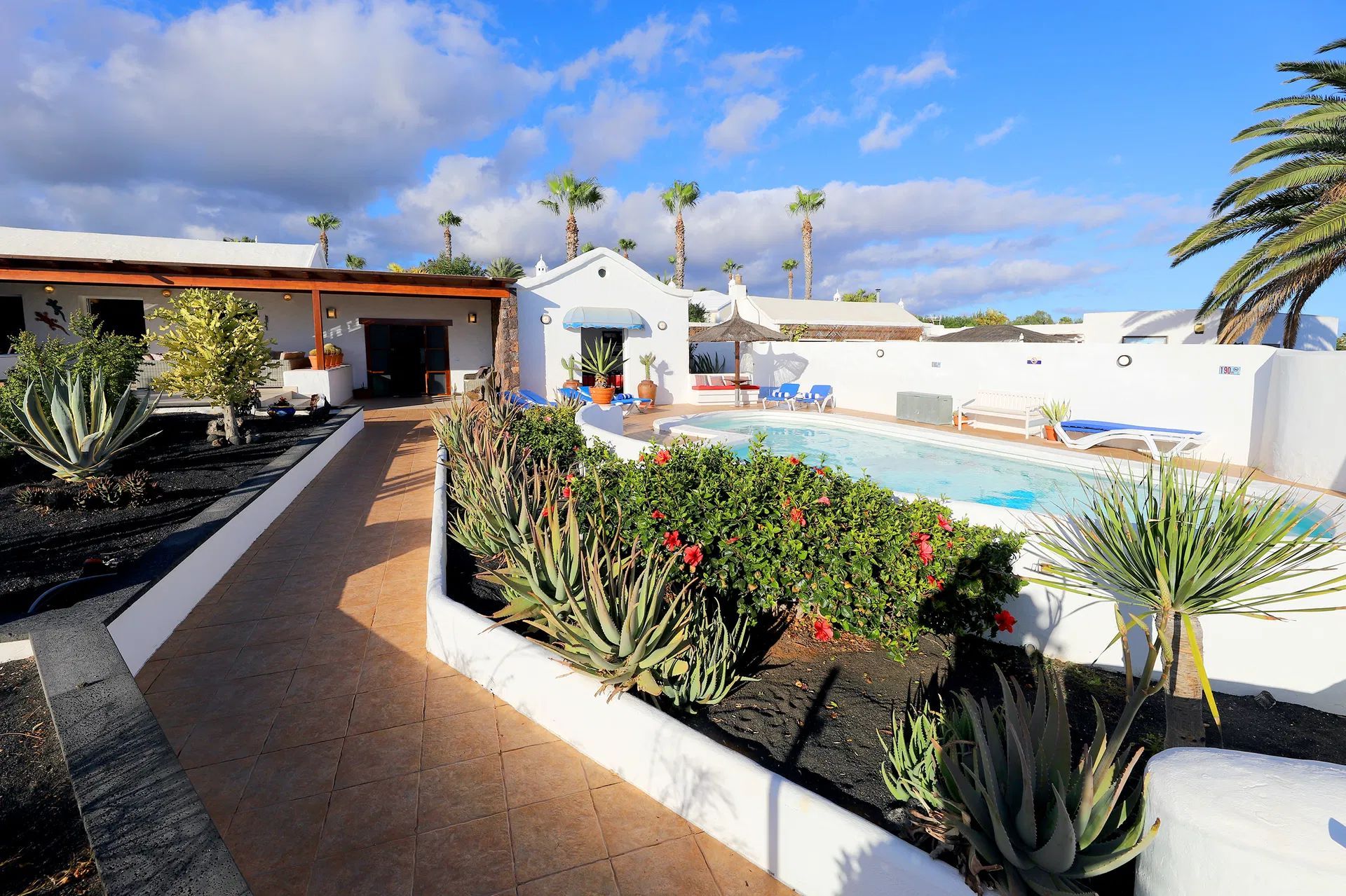 A pool area with white buildings, palm trees, and a pathway lined with plants under a blue sky. Villa Salvo | Los Mojones | Puerto del Carmen | Lanzarote.