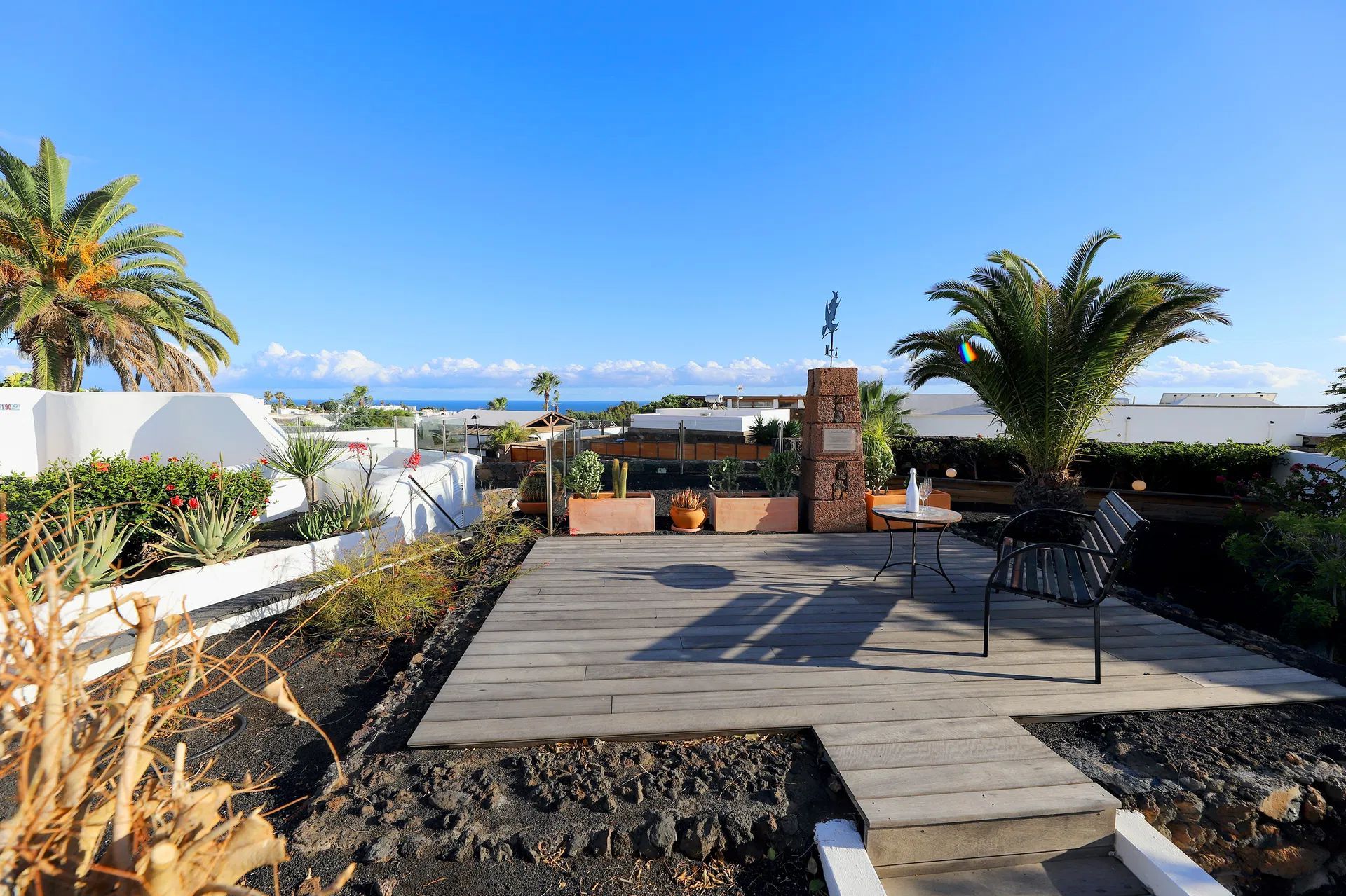 Wooden patio with table, chairs, plants, and a view of the ocean under a blue sky. Villa Salvo | Los Mojones | Puerto del Carmen | Lanzarote.