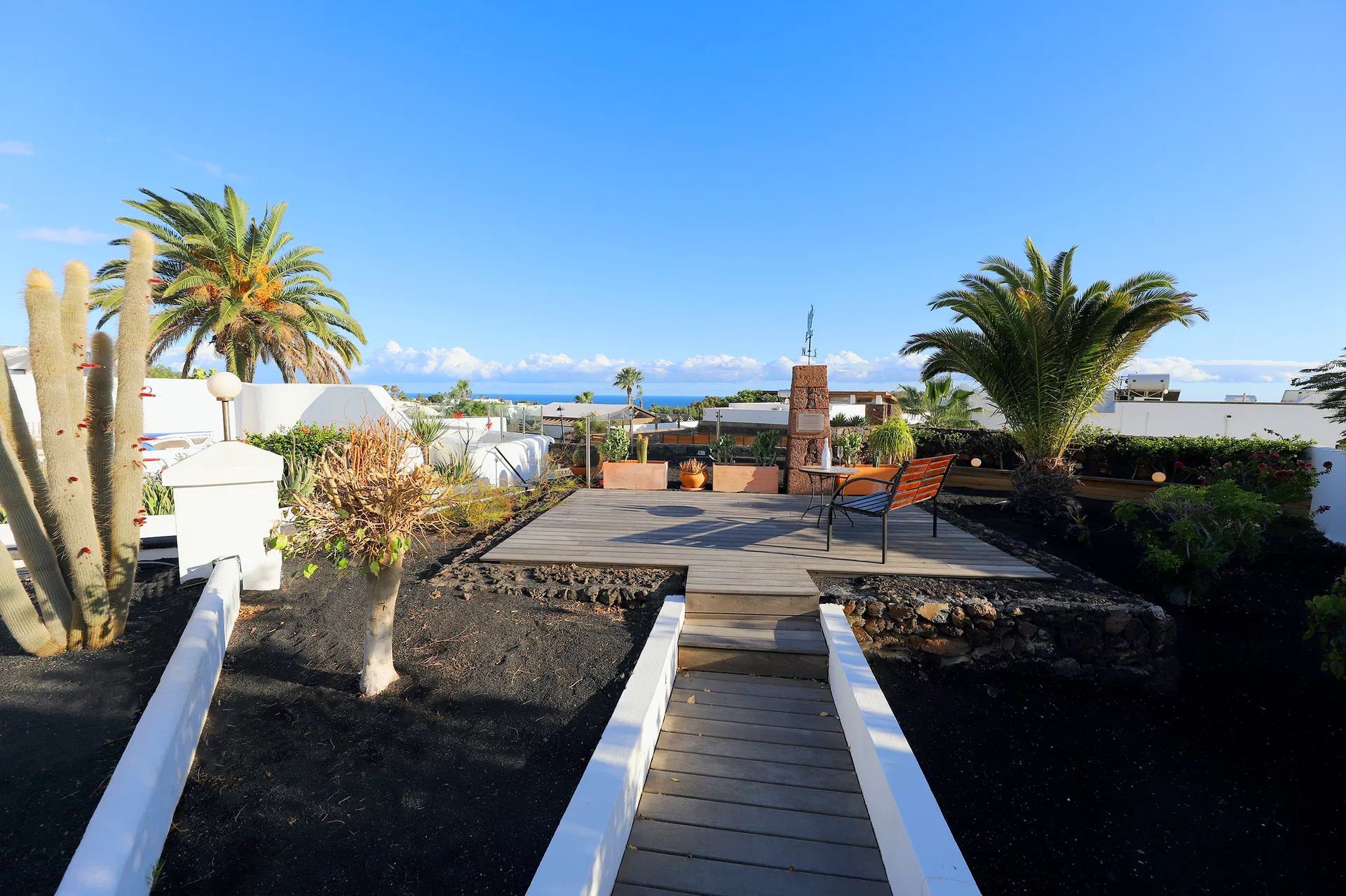 A patio with black lava rock and white walls, palm trees, and a view of the ocean under a blue sky. Villa Salvo | Los Mojones | Puerto del Carmen | Lanzarote.