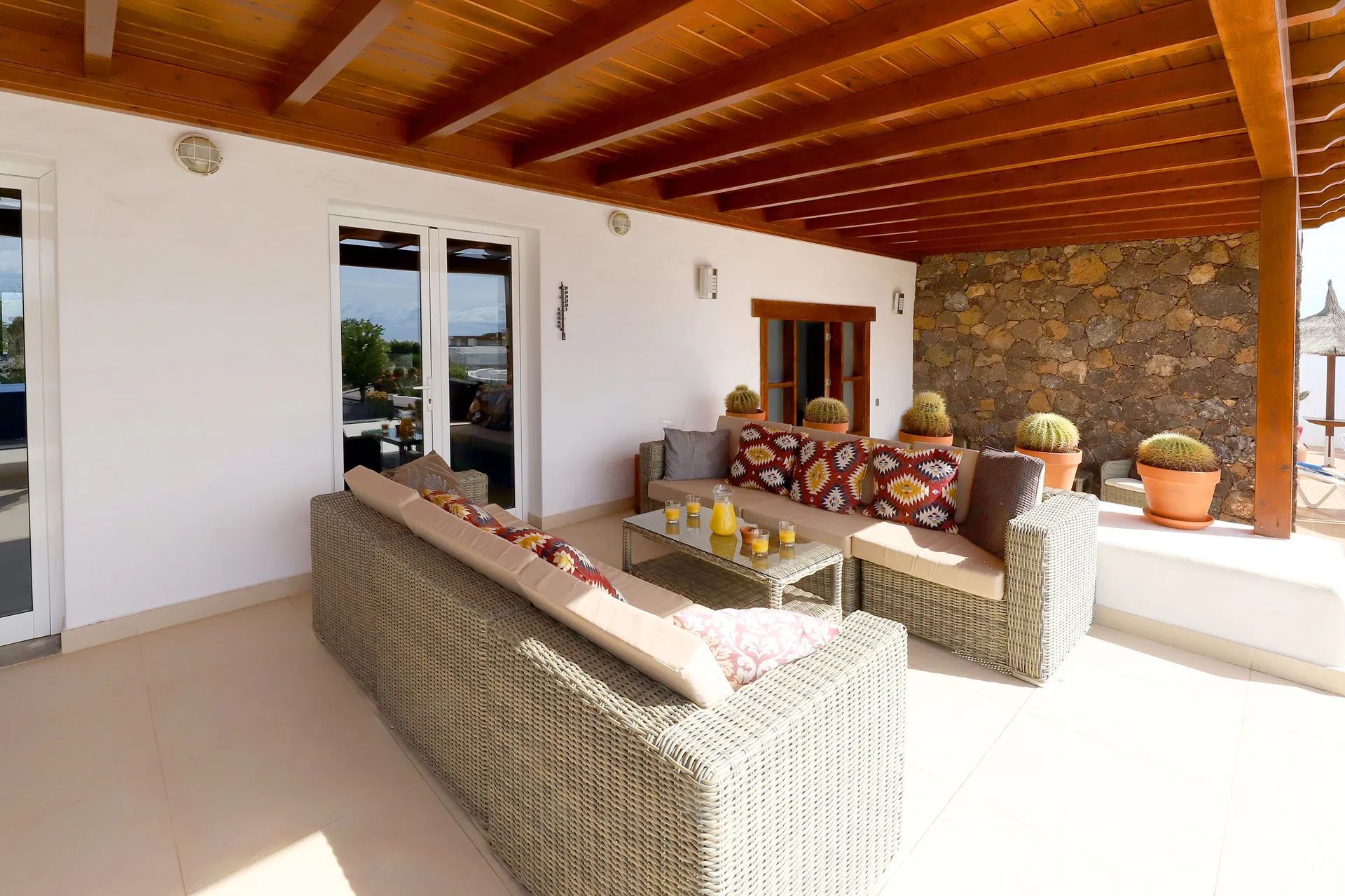 Outdoor patio with wicker furniture, stone wall, wooden ceiling, and potted plants. Villa Salvo | Los Mojones | Puerto del Carmen | Lanzarote.