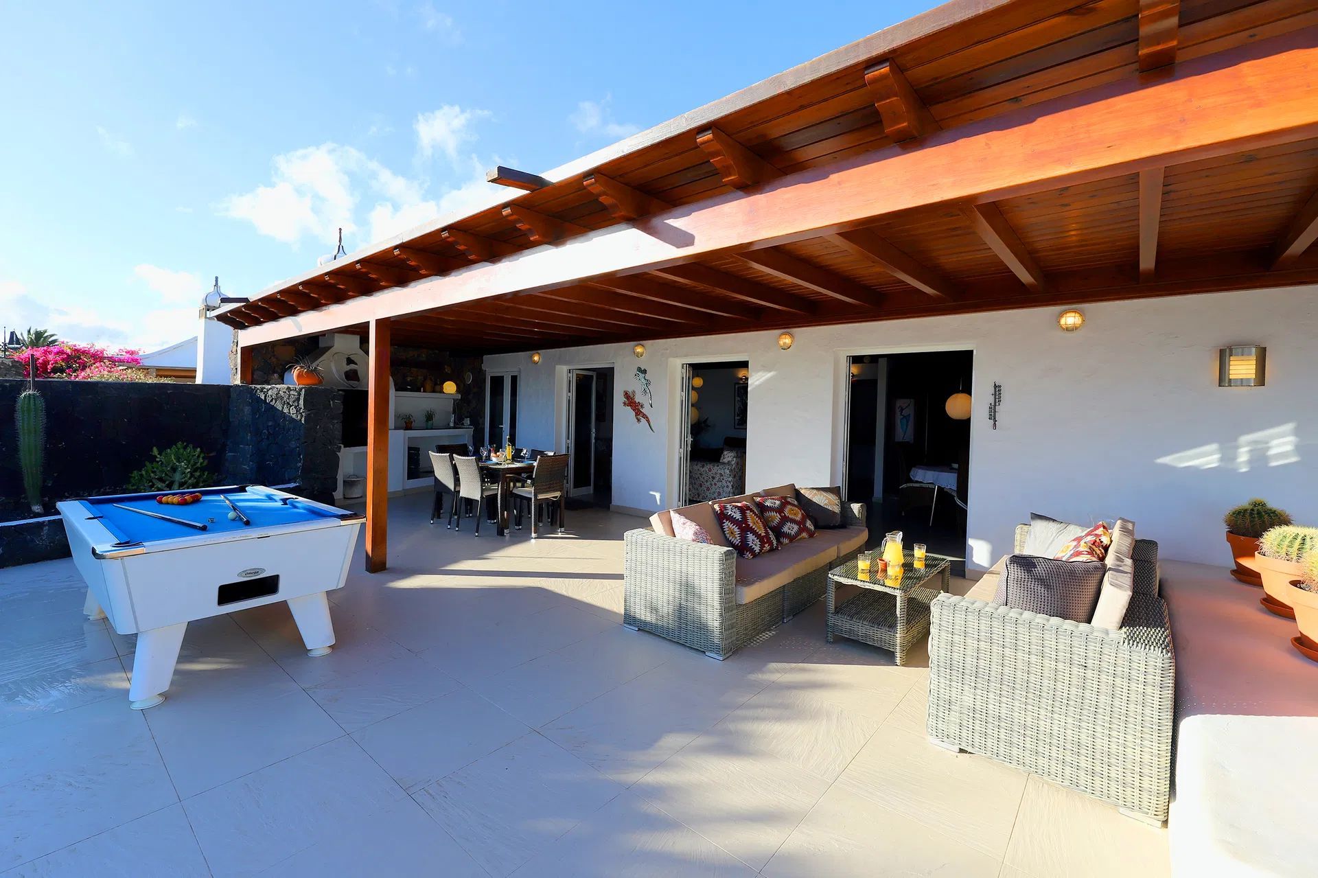 Patio with pool table, wicker furniture, dining table under a wooden pergola, and white walls. Villa Salvo | Los Mojones | Puerto del Carmen | Lanzarote.