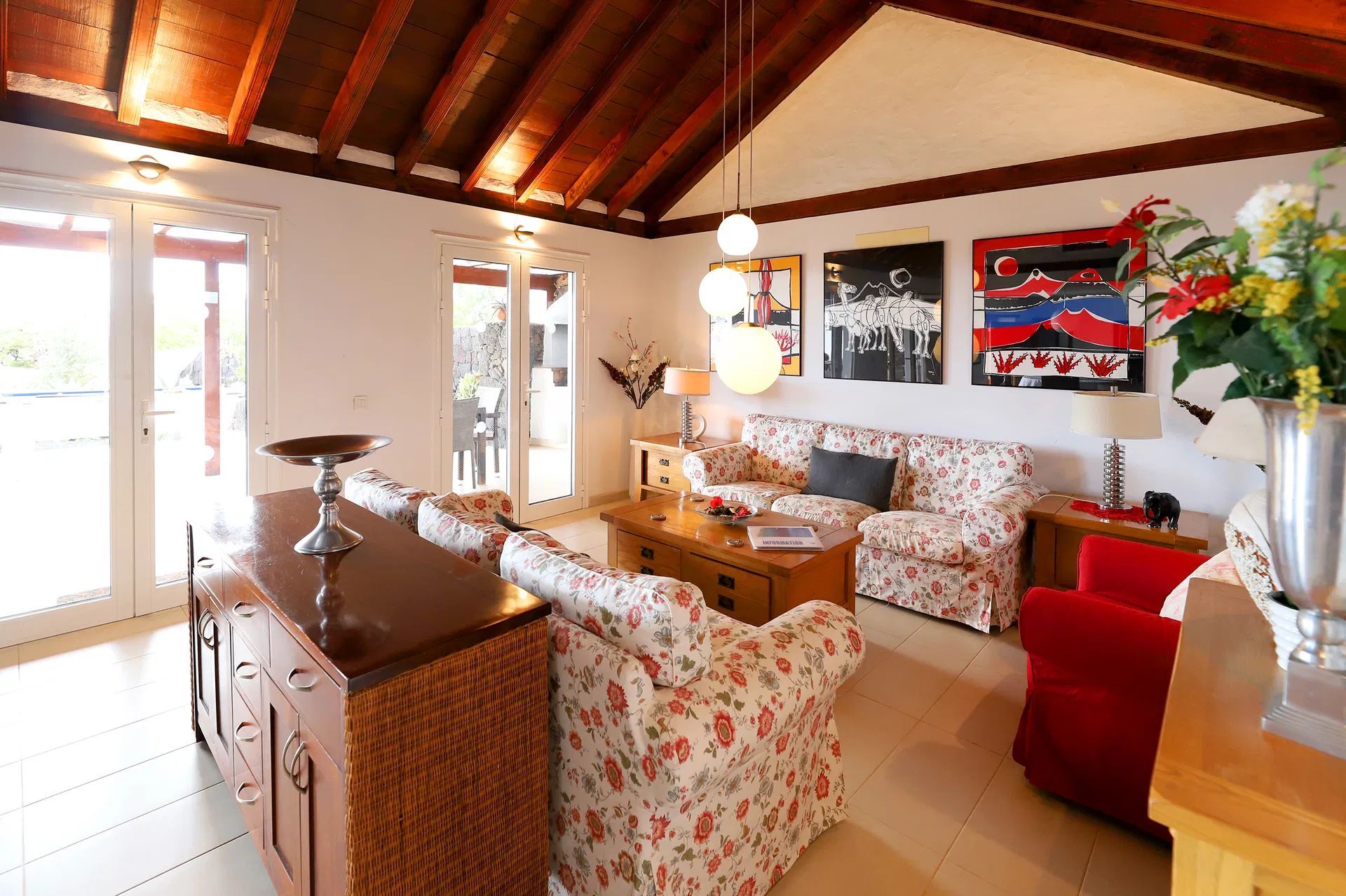 Living room with floral couches, red chair, art, and a wooden ceiling. Villa Salvo | Los Mojones | Puerto del Carmen | Lanzarote.