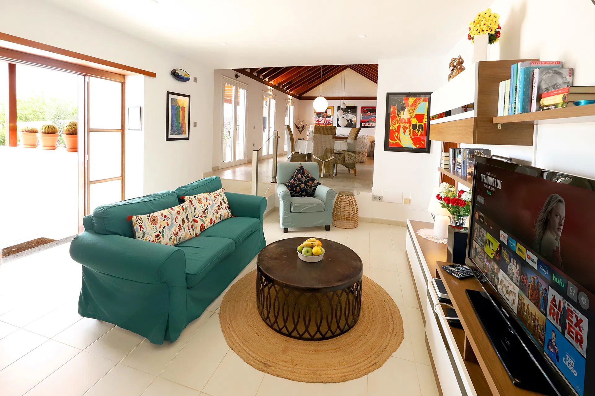 Living room with a green sofa, rug, and TV. Dining area visible beyond. Villa Salvo | Los Mojones | Puerto del Carmen | Lanzarote.