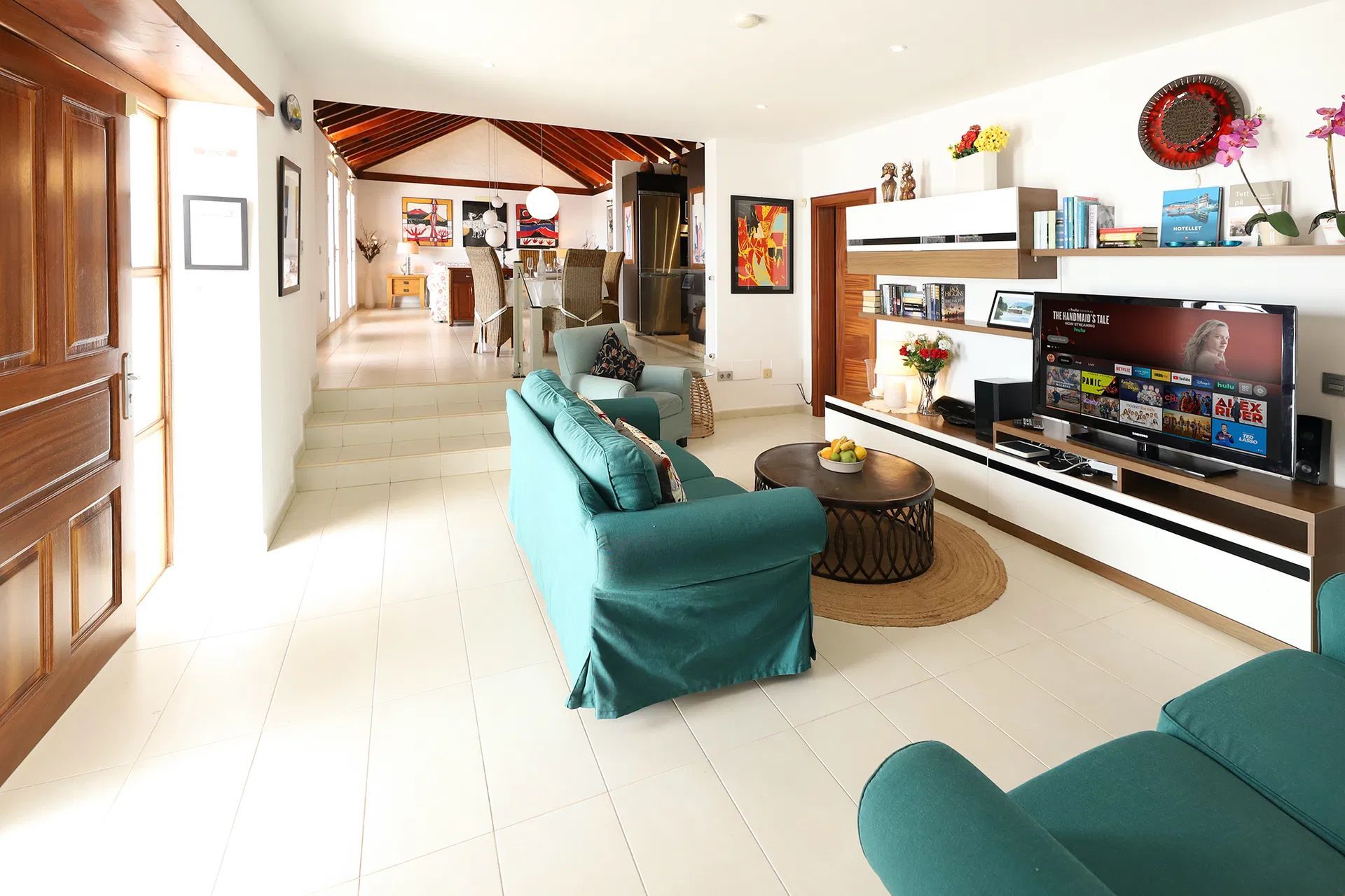 Bright living room with teal sofa, TV, and artwork; a doorway leads to another space. Villa Salvo | Los Mojones | Puerto del Carmen | Lanzarote.