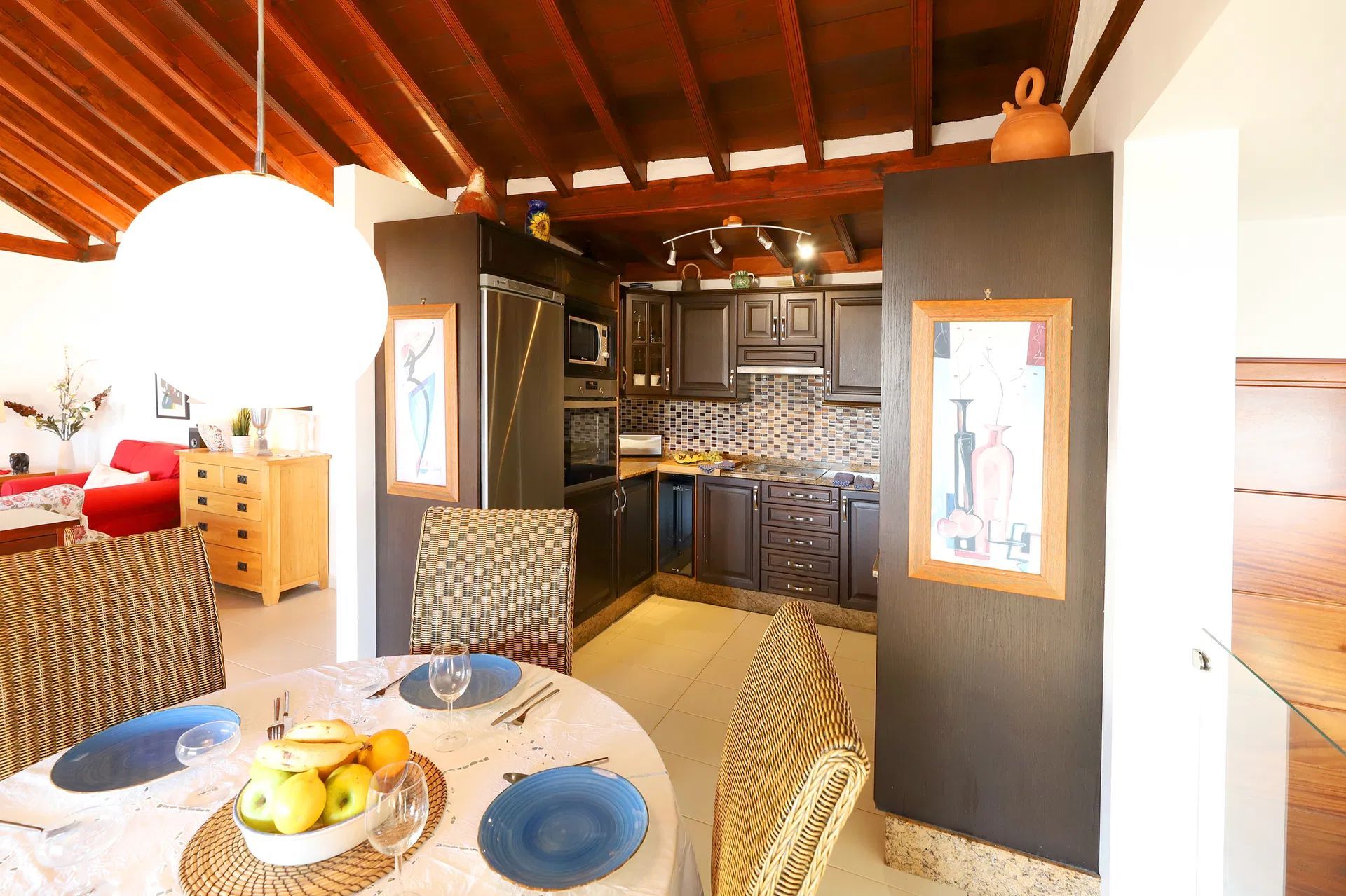 Dining room with kitchen visible: round table, blue plates, fruit bowl, dark cabinets, wooden beams. Villa Salvo | Los Mojones | Puerto del Carmen | Lanzarote.