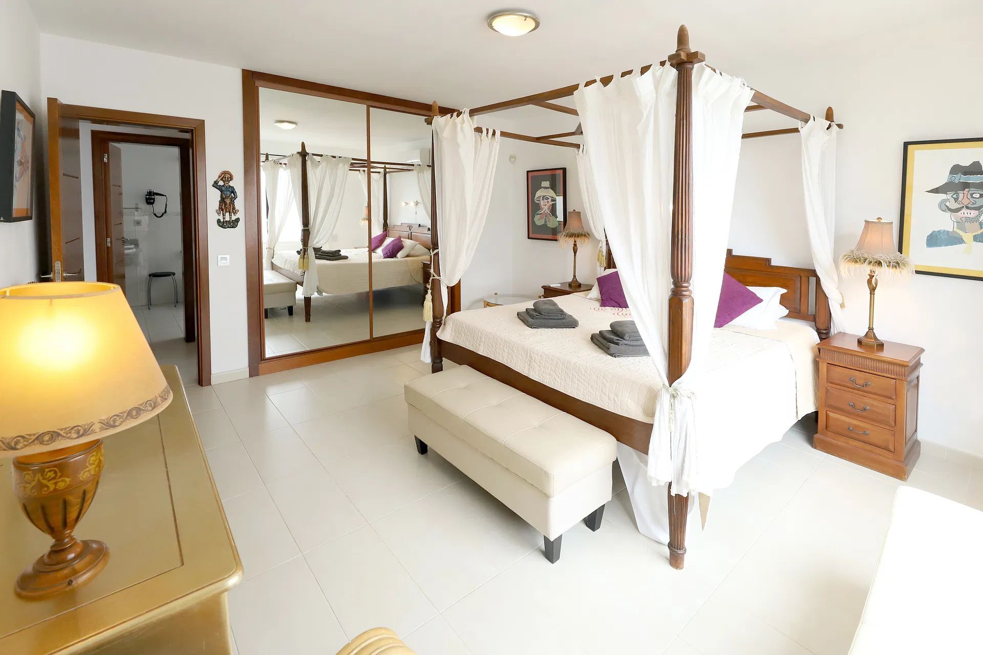Bedroom with four-poster bed, mirrored closet, side table, lamp, and door to a bathroom. Villa Salvo | Los Mojones | Puerto del Carmen | Lanzarote.