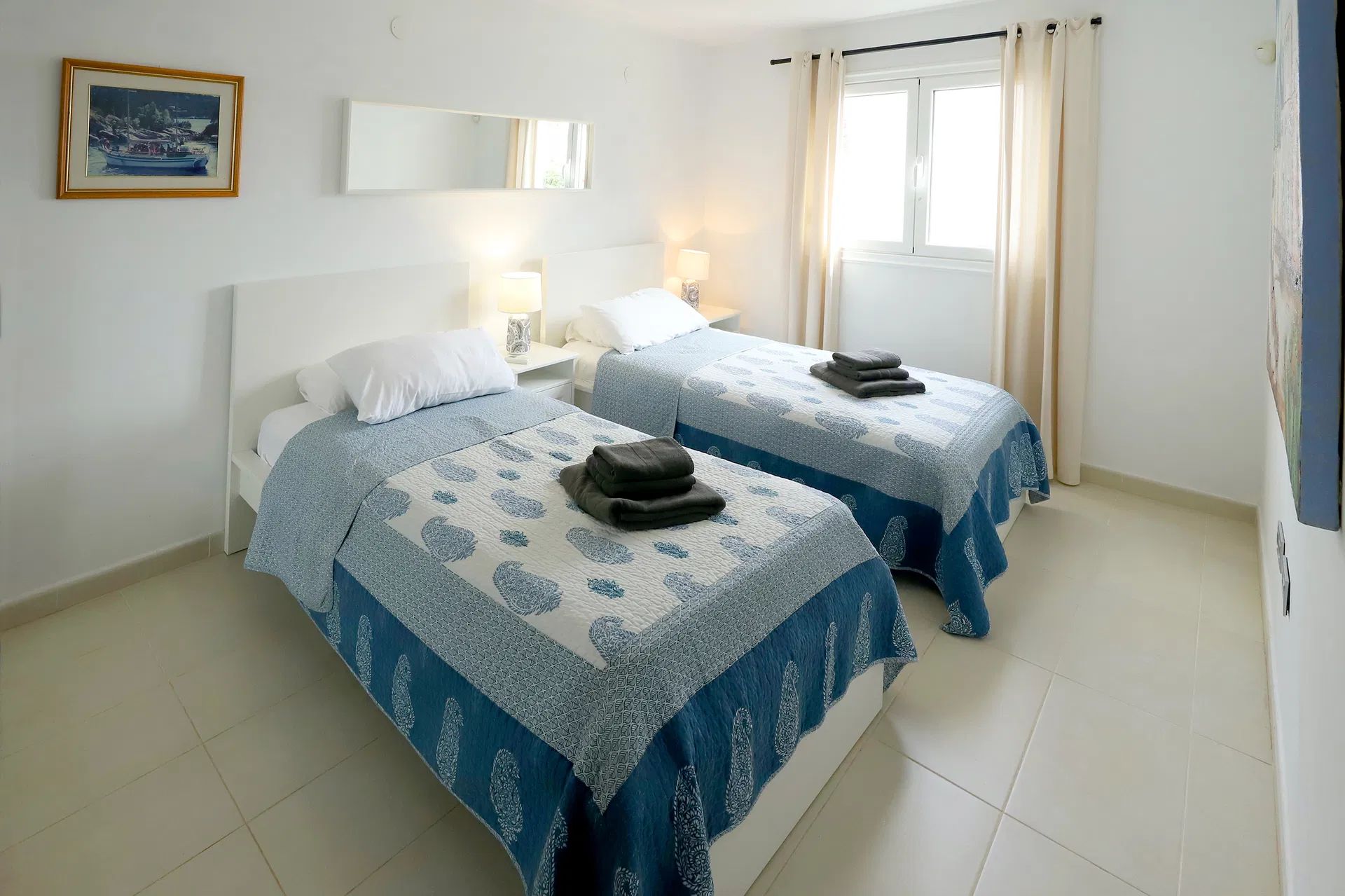 Two twin beds in a bright bedroom, each with blue patterned quilt, white headboard, and window with curtains. Villa Salvo | Los Mojones | Puerto del Carmen | Lanzarote.