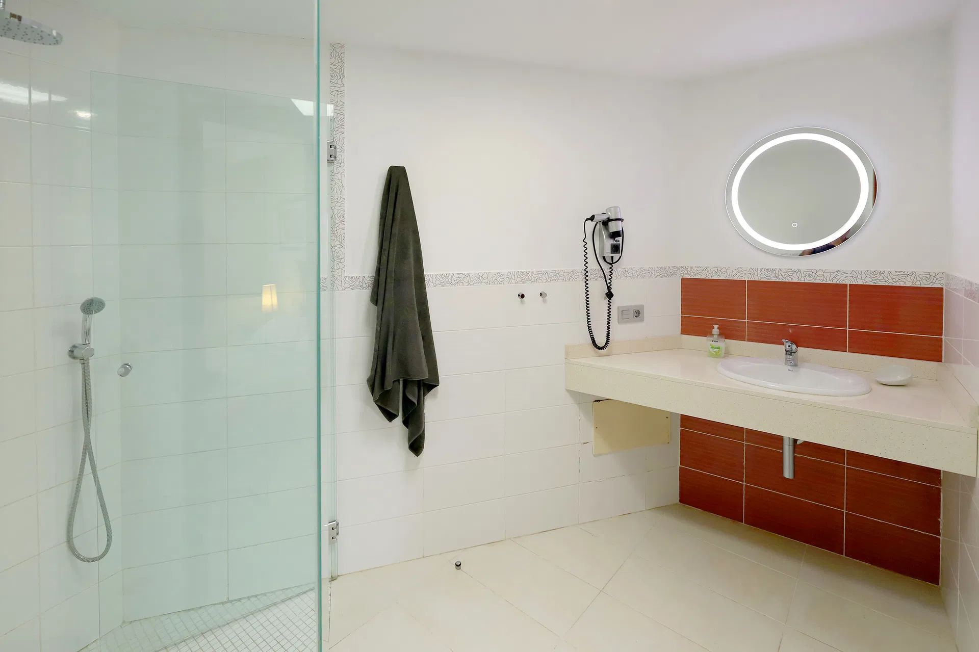 Bathroom with glass shower, white walls, red tile, and a lit round mirror. Villa Salvo | Los Mojones | Puerto del Carmen | Lanzarote.