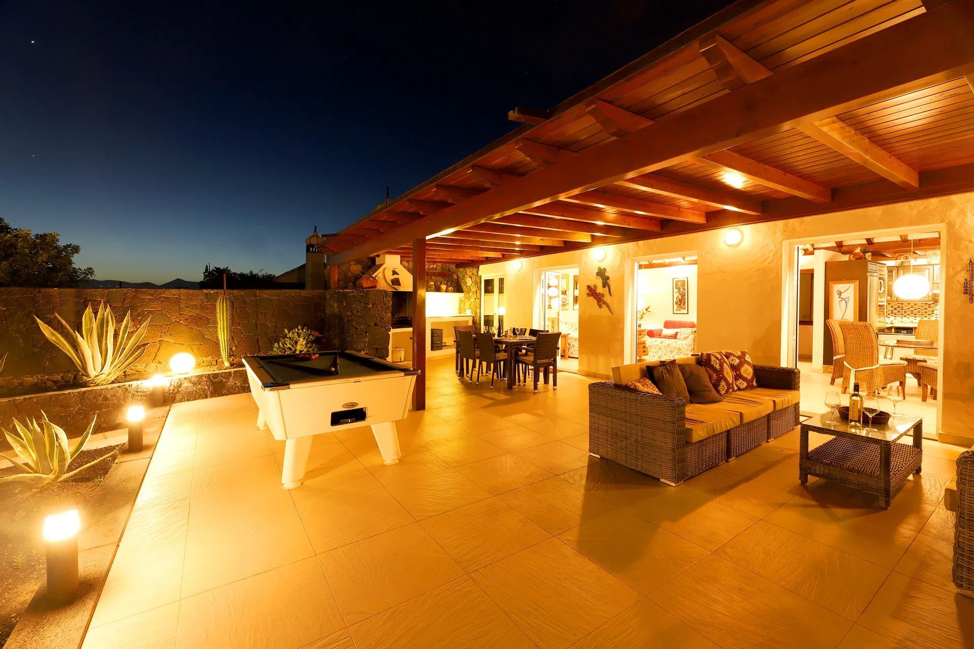 Outdoor patio at dusk with a pool table, seating area, and dining table under a wooden pergola. Villa Salvo | Los Mojones | Puerto del Carmen | Lanzarote.
