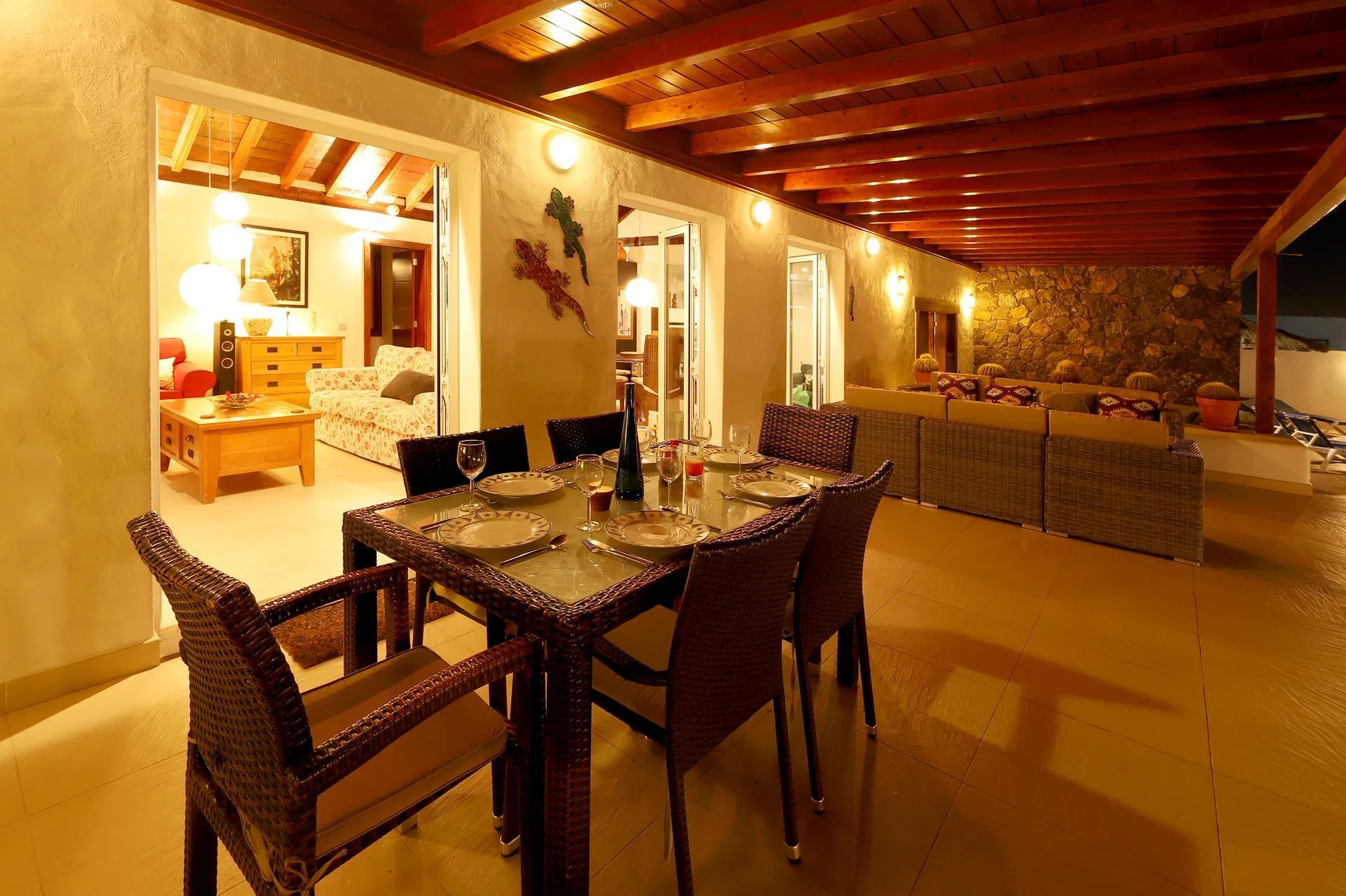 Outdoor dining area with table set for a meal, overlooking an interior room, at night. Villa Salvo | Los Mojones | Puerto del Carmen | Lanzarote.